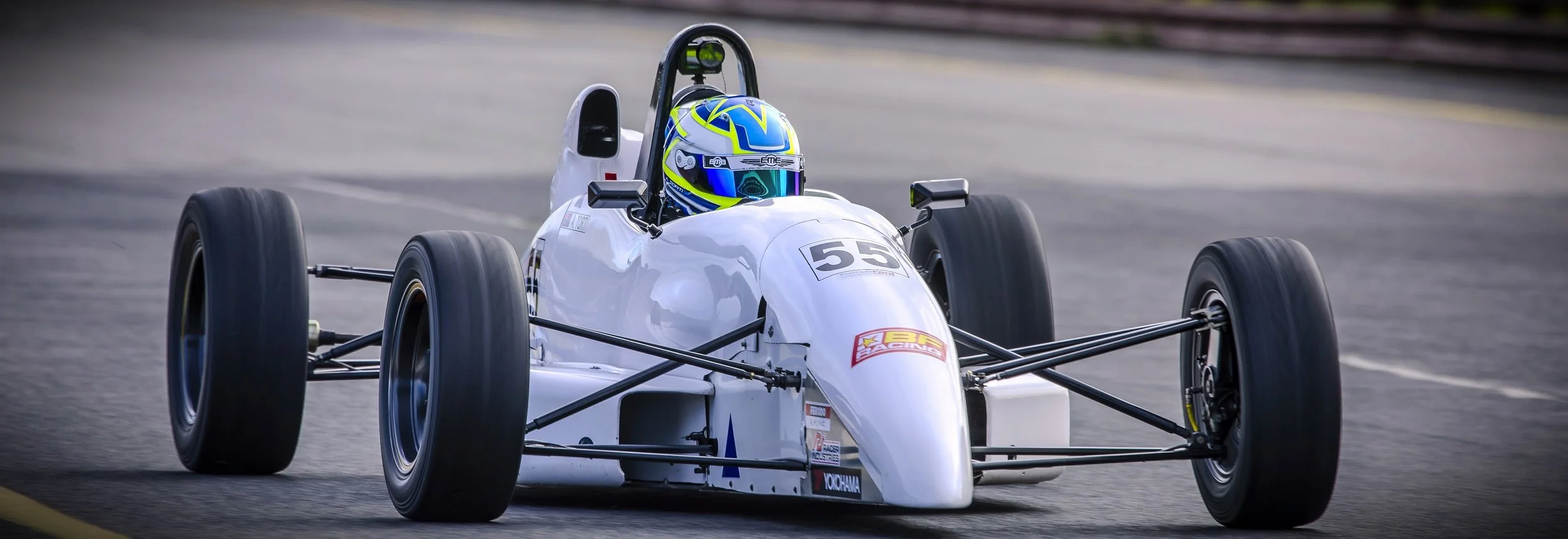 A white vintage race car with number 55, driven by a driver in a blue and yellow helmet, on a racetrack.