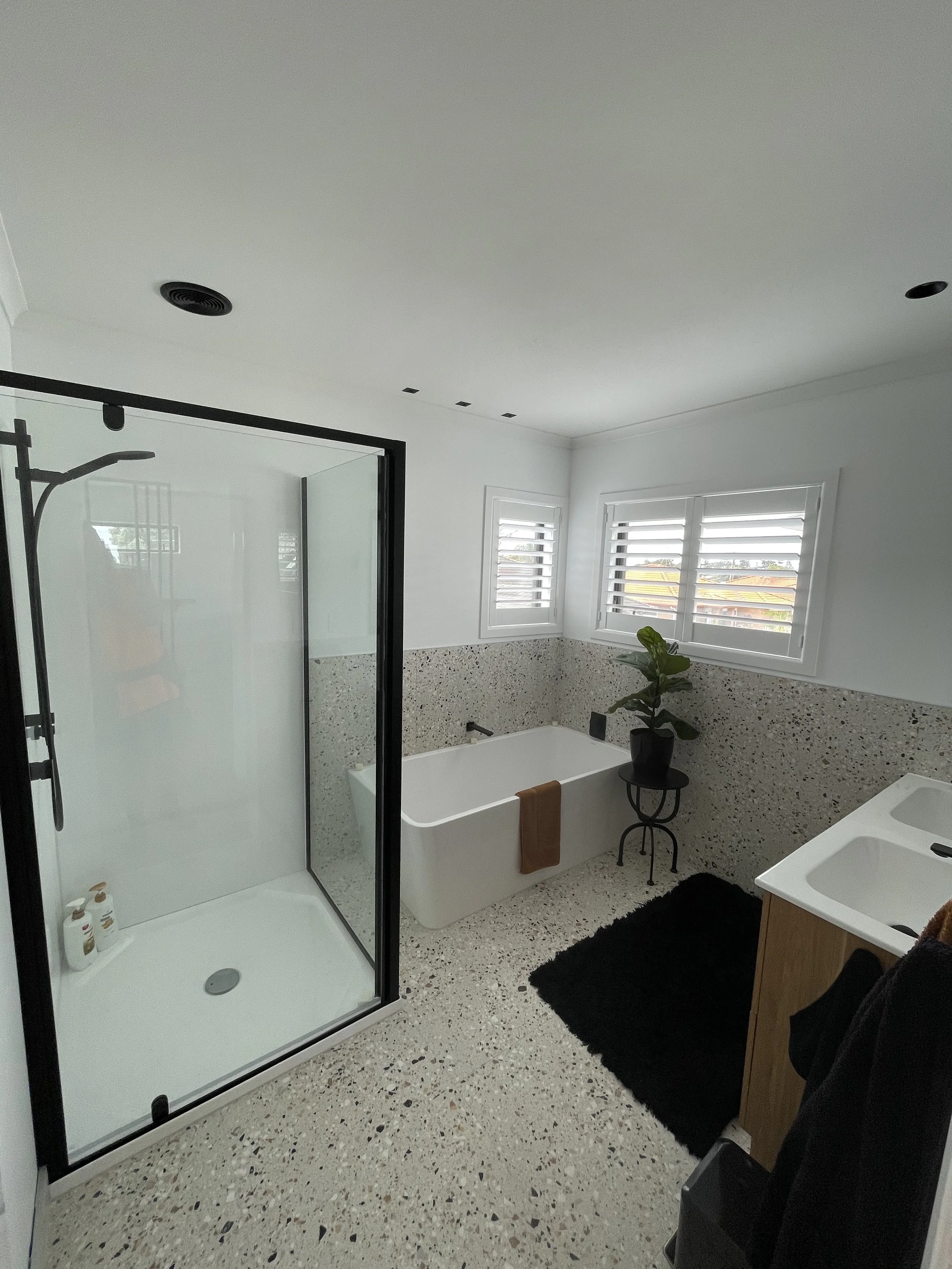Modern bathroom with a shower, a bathtub, a black potted plant, two windows with white shutters, black bath mat, and a vanity with two sinks.