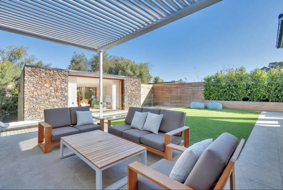 Modern outdoor patio with seating, stone and wooden house, green lawn, and blue sky.