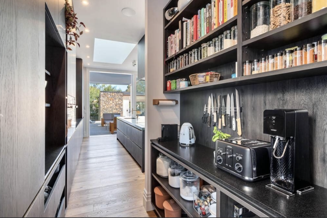 Modern kitchen with black shelves, spices, knives, toaster, coffee machine, and glass jars, leading to a bright outdoor patio.