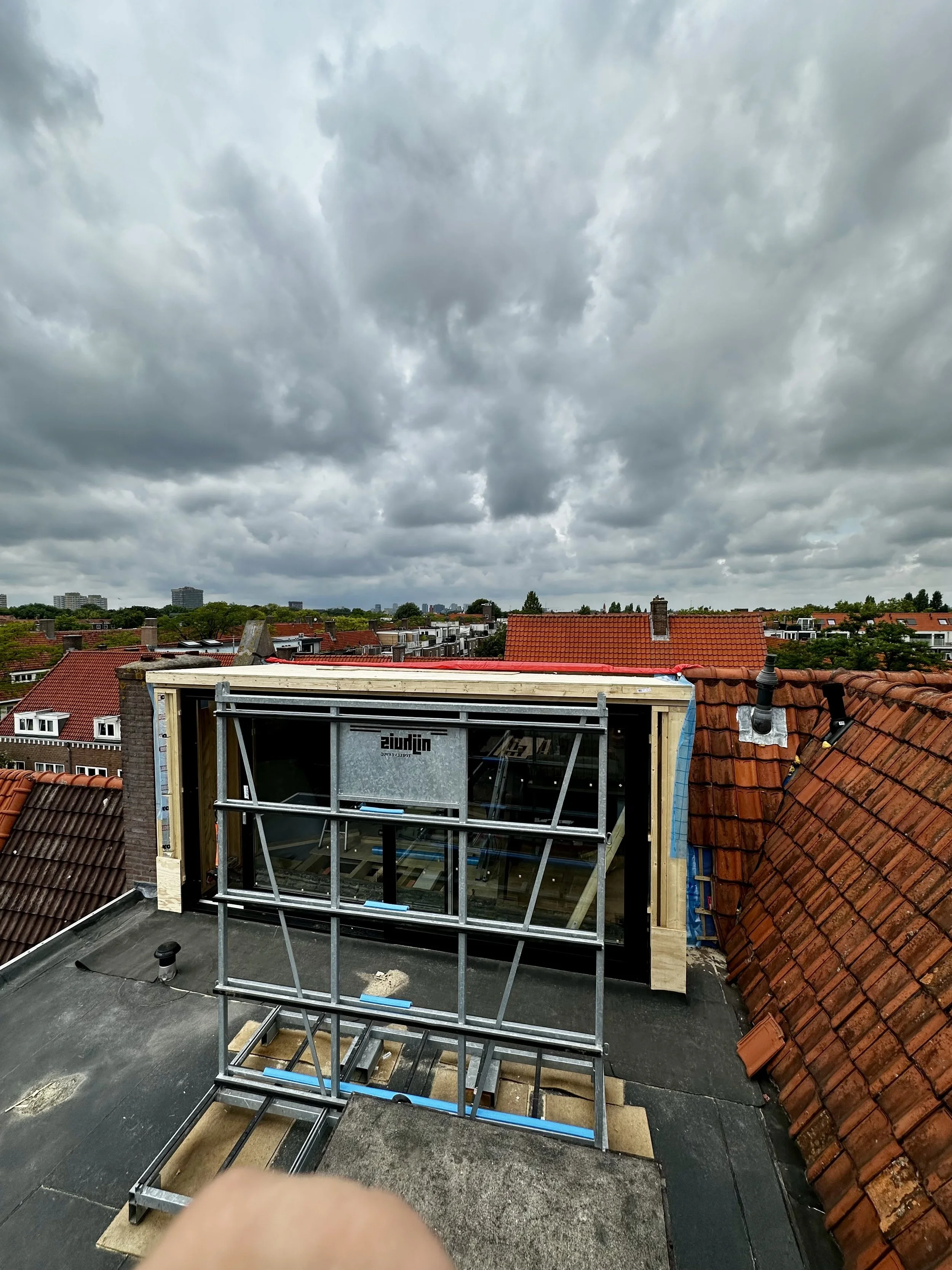 View of a rooftop under construction with a window frame and scaffolding, surrounded by red-tiled roofs and dark cloudy sky.