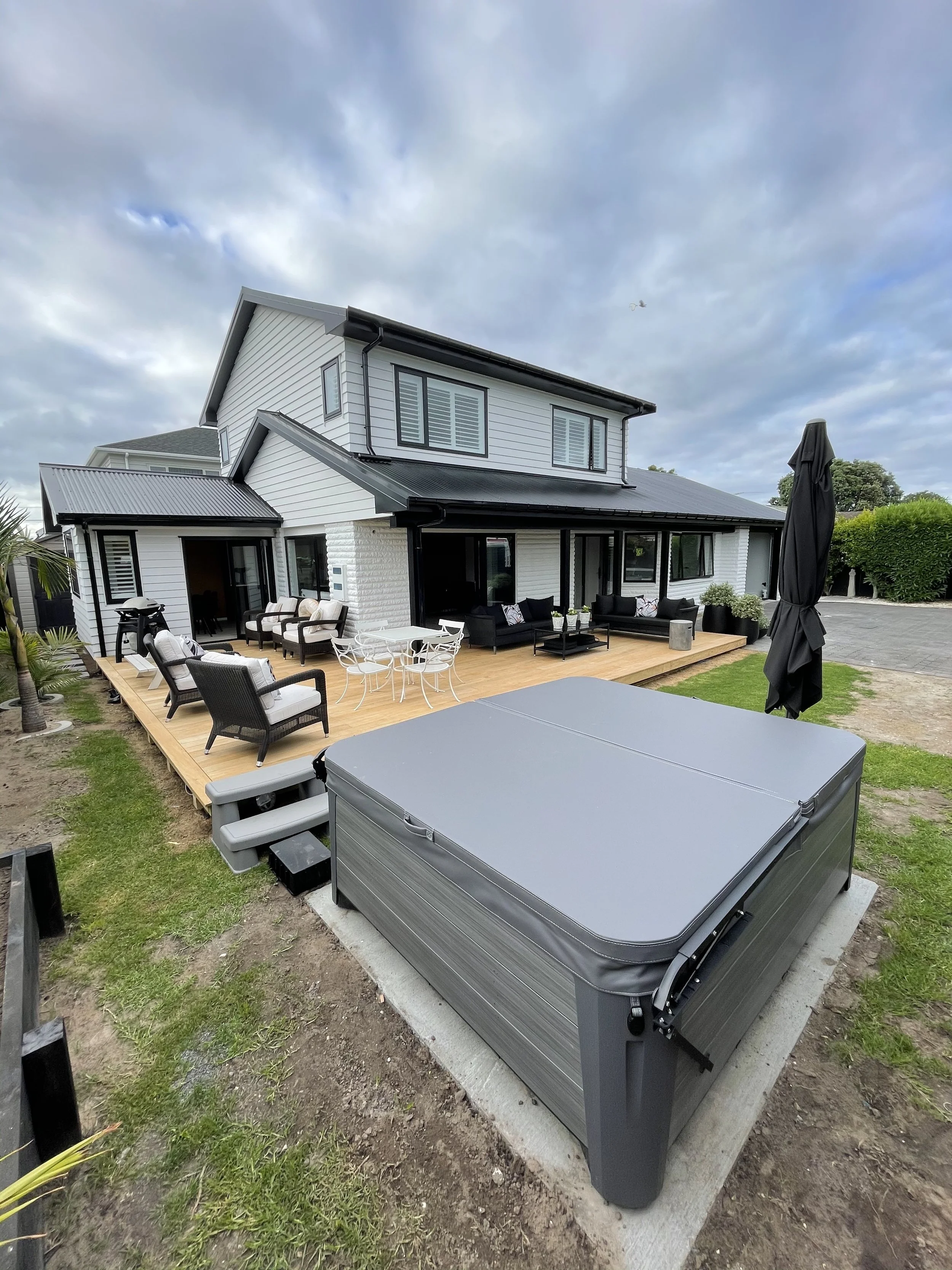 Modern two-story house with white siding and black trim, outdoor patio with black and white furniture, hot tub on grass, cloudy sky.