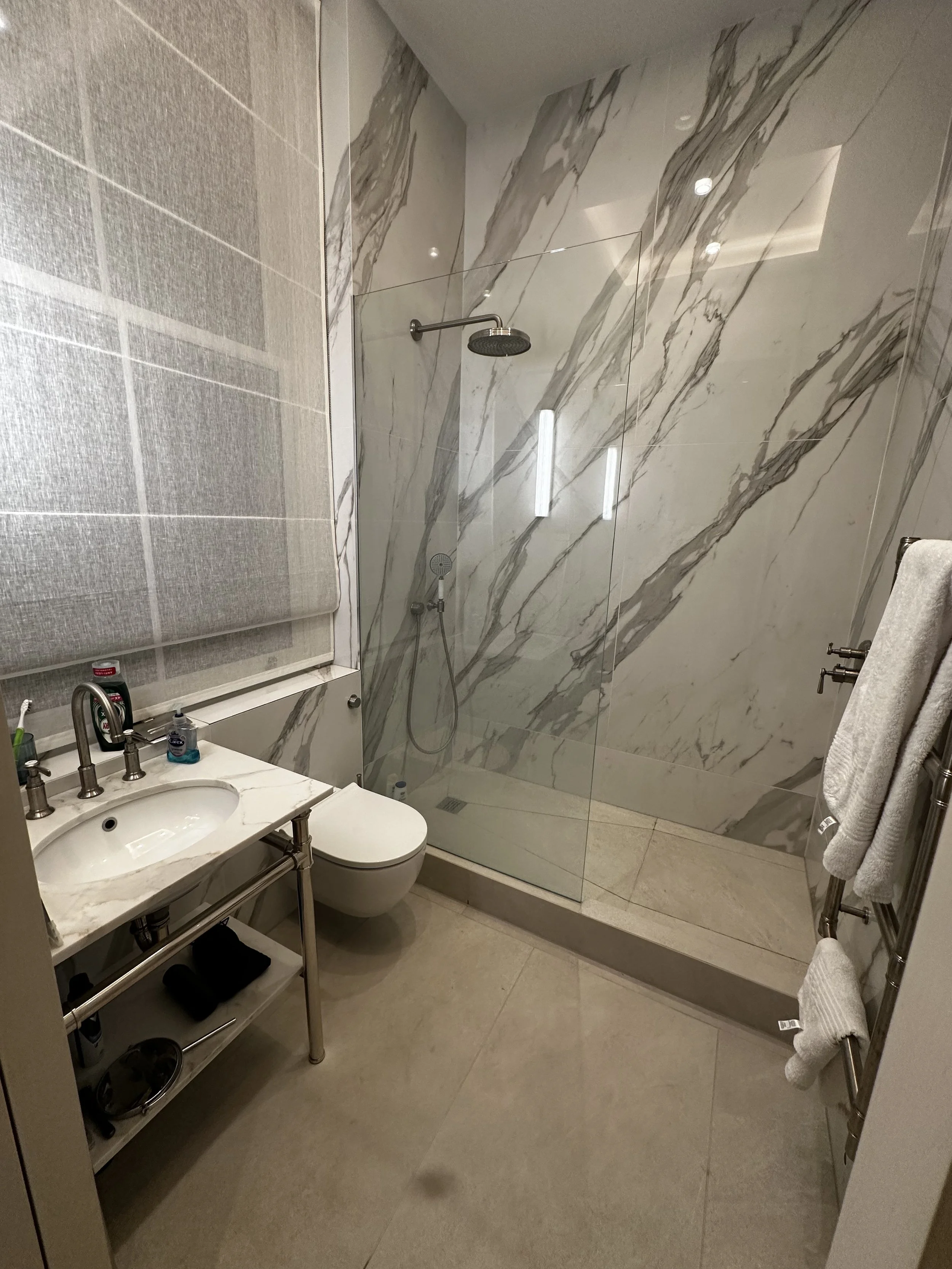 Modern bathroom with a walk-in shower featuring marble walls and a glass enclosure, a toilet, a small marble countertop sink with chrome fixtures, and a heated towel rack with white towels.