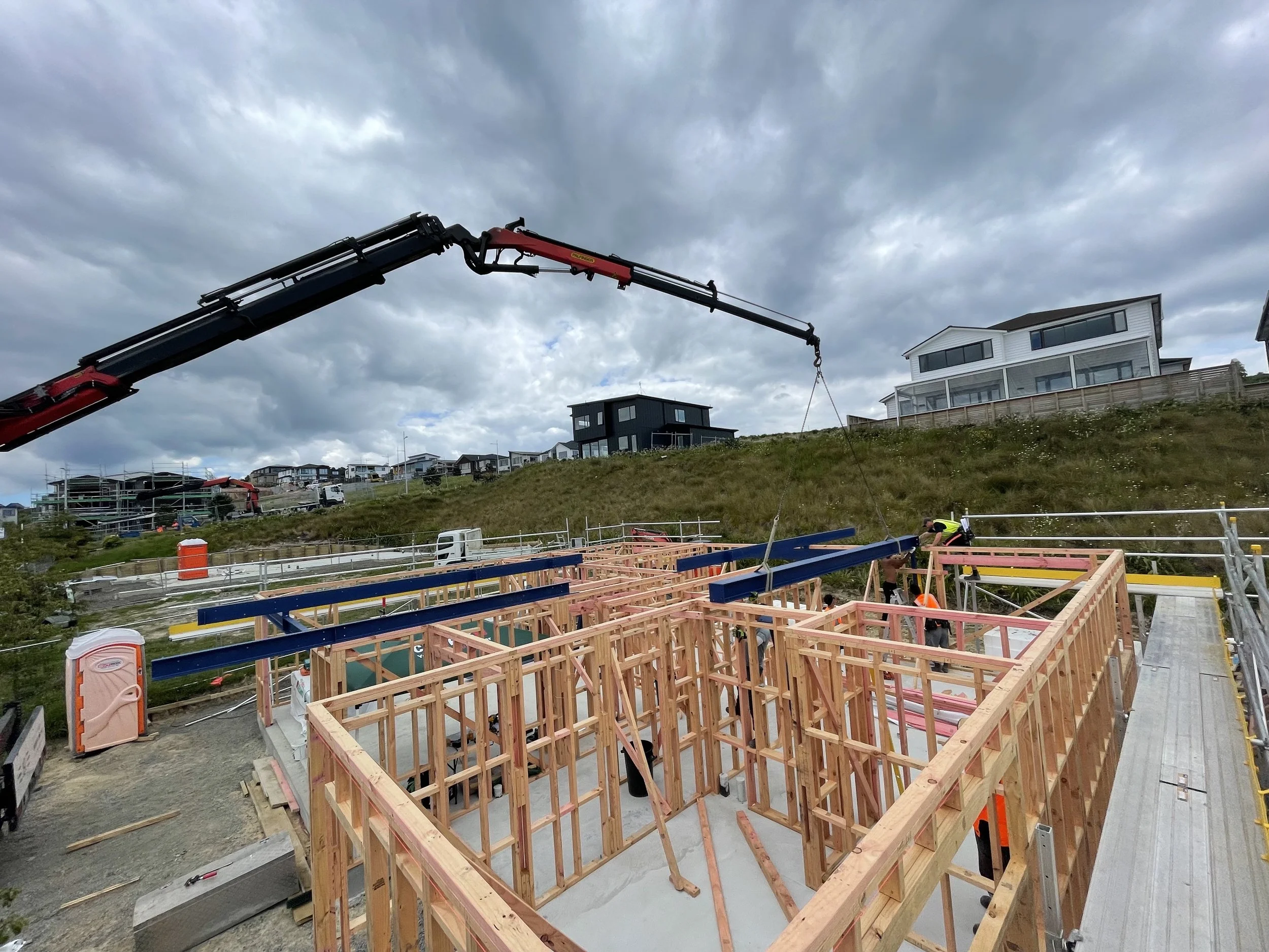 Construction site with wooden framing, workers assembling, and a crane lifting a steel beam against a cloudy sky, residential houses on a hillside in the background.
