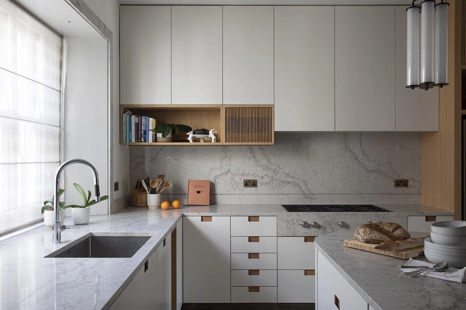 A modern kitchen with white cabinetry, marble countertops, and a marble backsplash. There is a sink with a silver faucet by a window with sheer curtains. On the counter, there are two oranges, a pink book, a white vase with plants, and various cooking utensils. A loaf of bread is on a wooden cutting board, and there are bowls and utensils nearby.