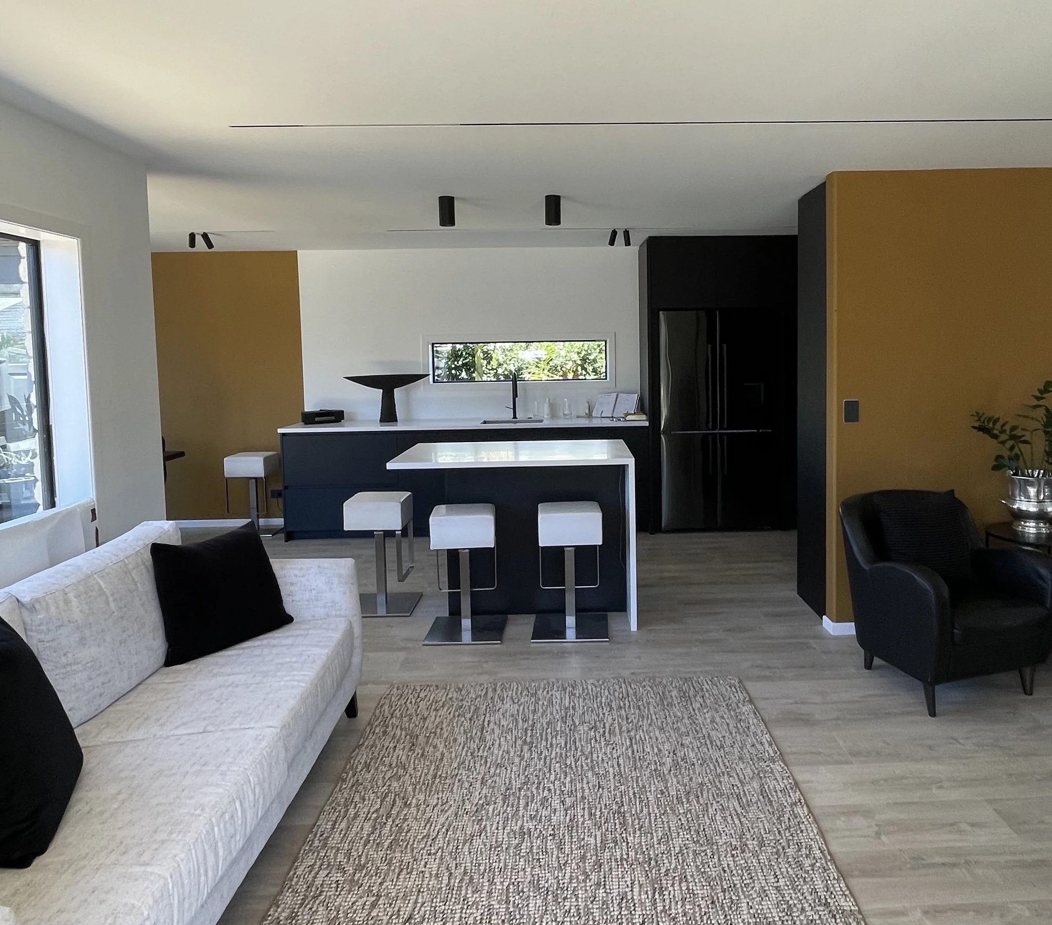Modern open-concept living room and kitchen with black and white furniture, gold accent wall, hardwood floor, large window, and minimalist decor.