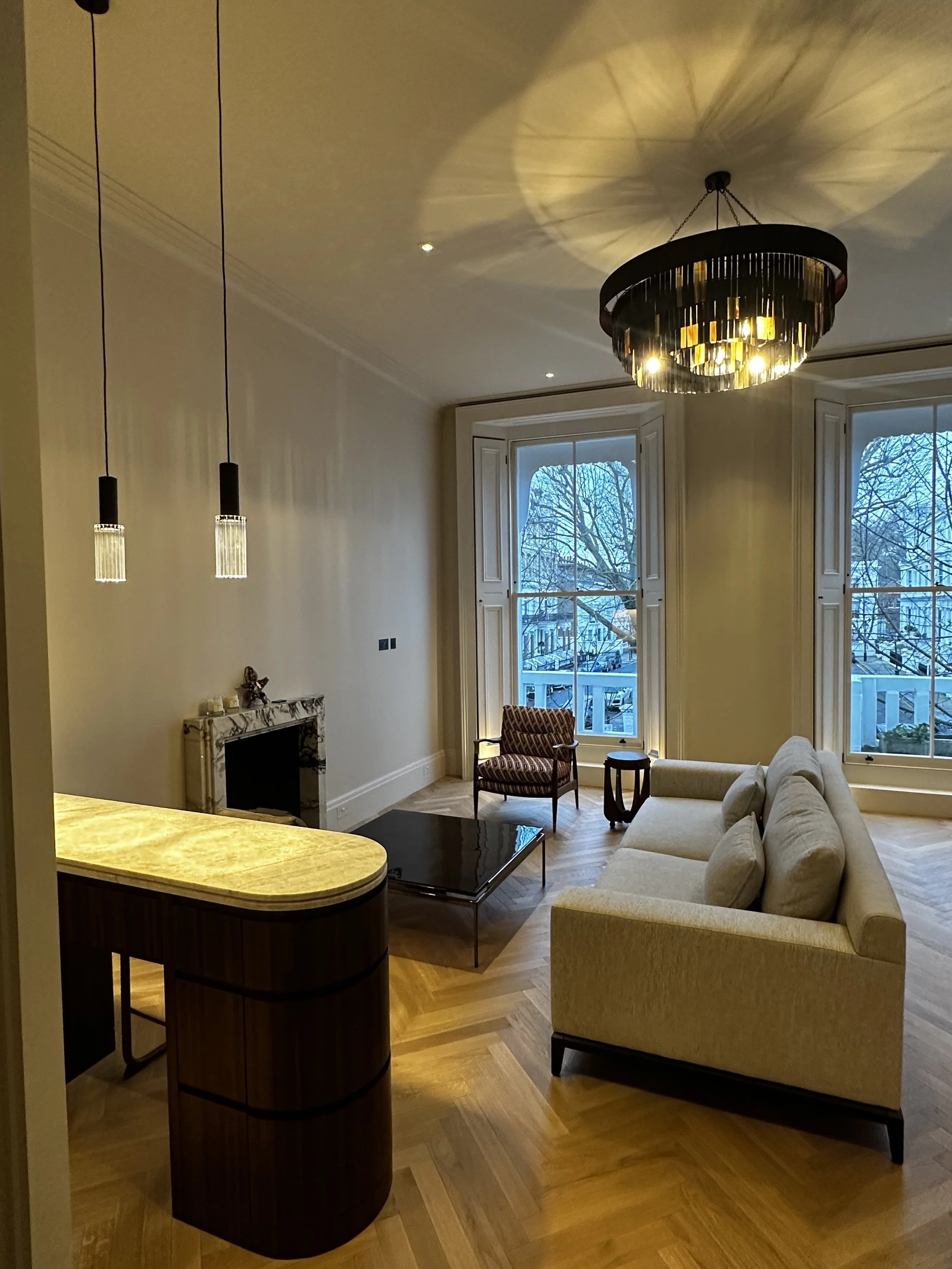 Living room with large windows, beige sofa, patterned armchair, wood floor, black coffee table, marble fireplace, chandelier, and hanging pendant lights.