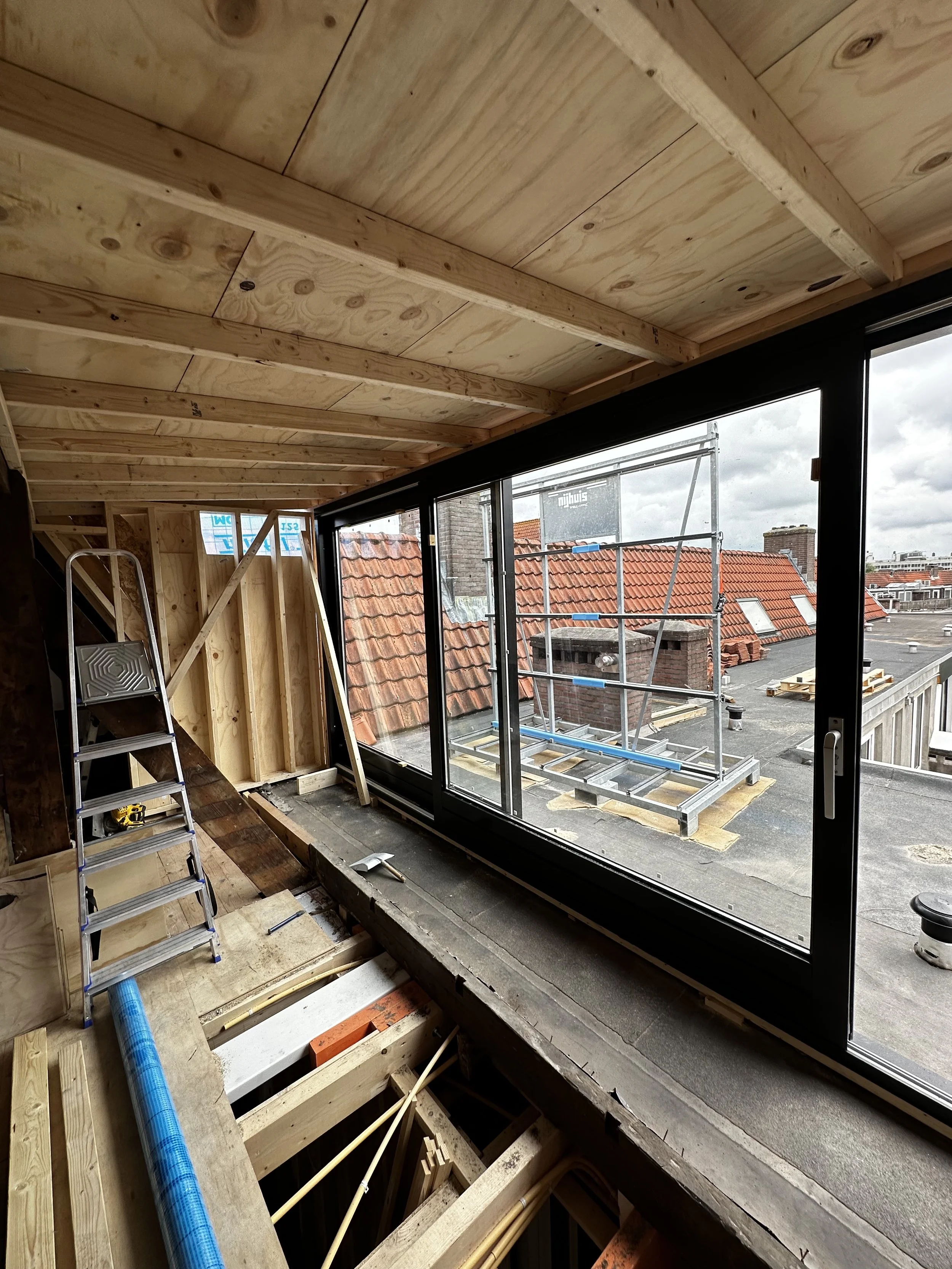 Interior of a building under construction with a large sliding glass door, wooden framing, and a ladder, overlooking rooftops and a cloudy sky outside.