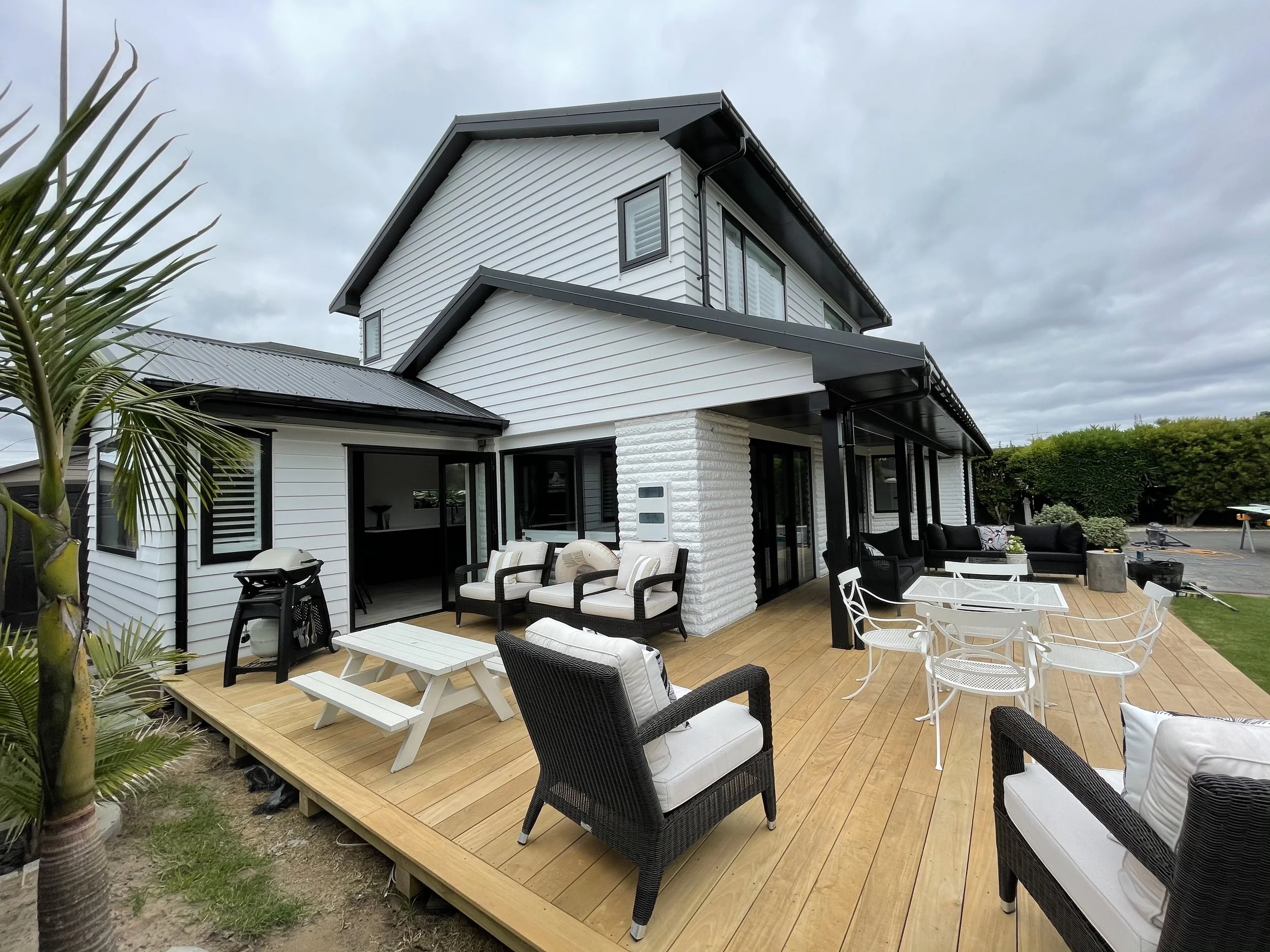 Modern house with a large wooden patio, outdoor furniture including chairs, tables, and couches, and a grill, under an overcast sky.