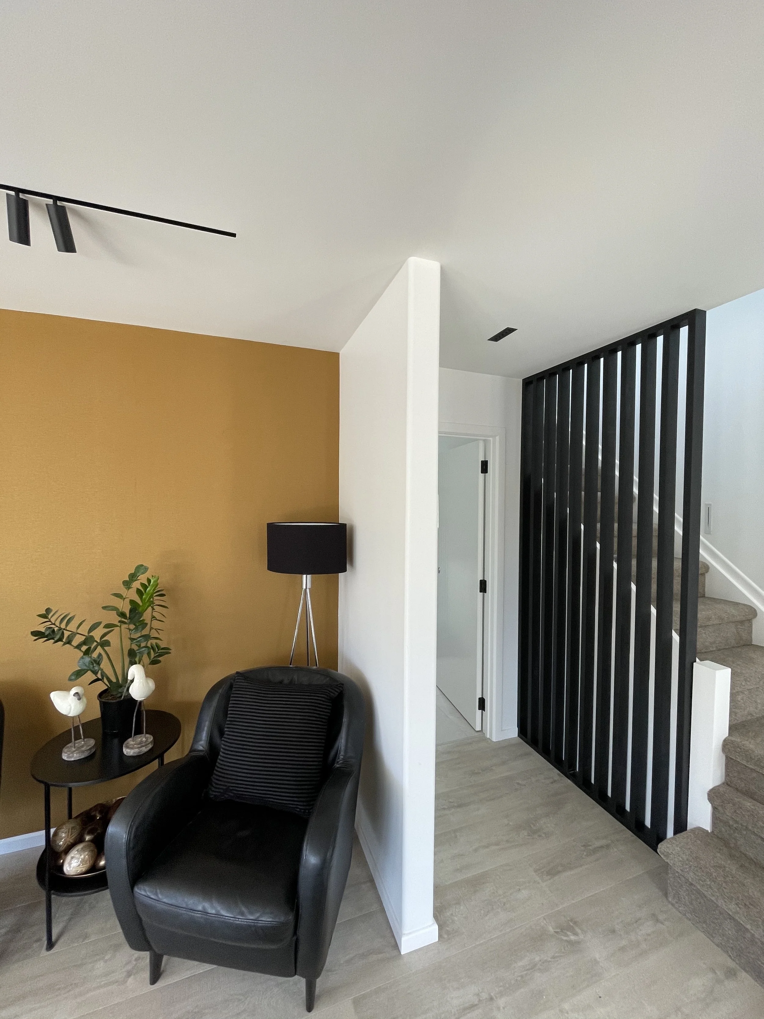 A modern living room corner with a black leather armchair, black throw pillow, small round black side table with decorative objects, potted plant, black floor lamp, white wall, yellow accent wall, black vertical slat room divider, carpeted staircase, and light-colored wood flooring.