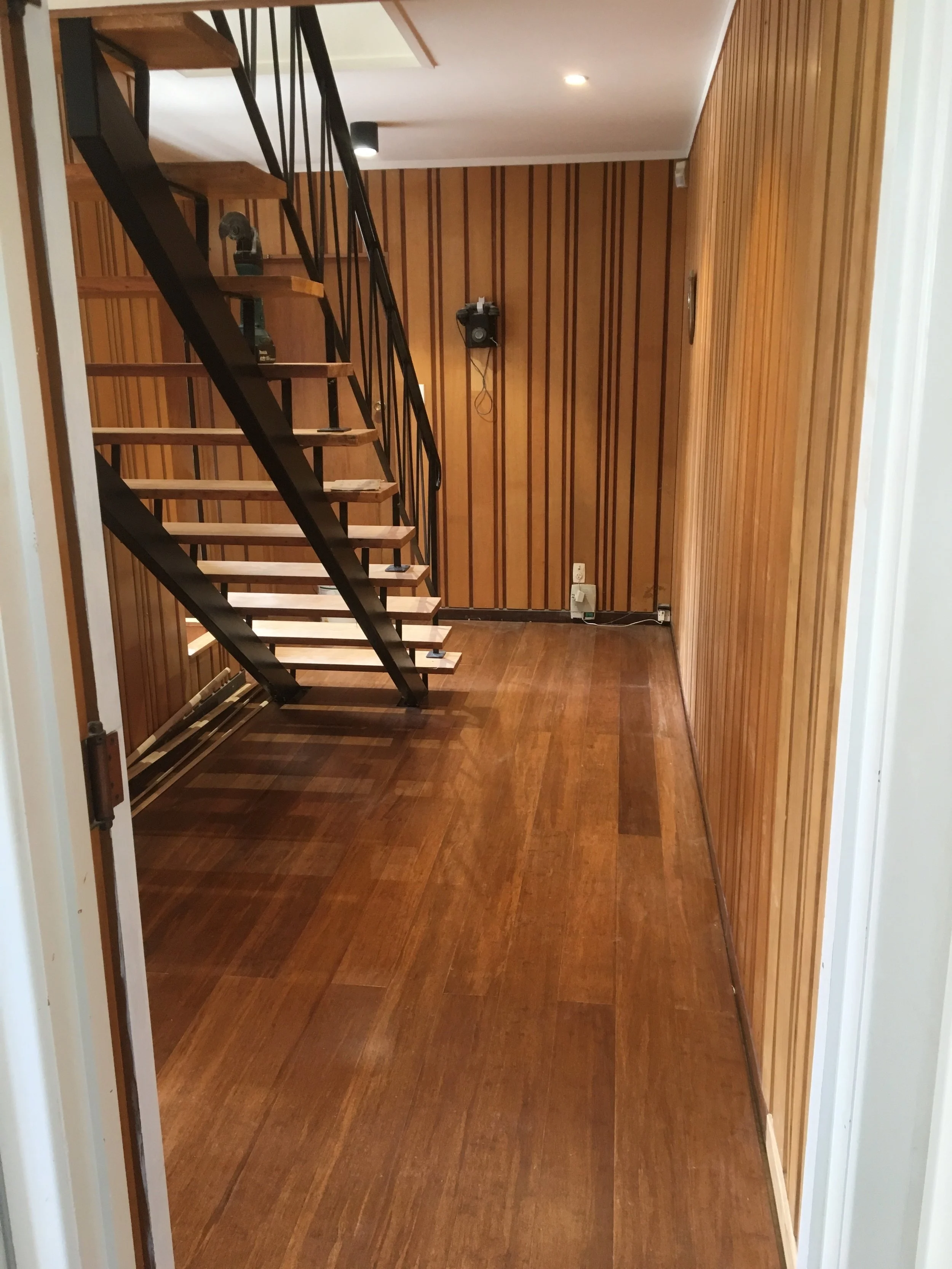 Empty room with wood-paneled walls and wooden floor, with a black metal staircase leading upstairs, and a wall-mounted speaker on the back wall.