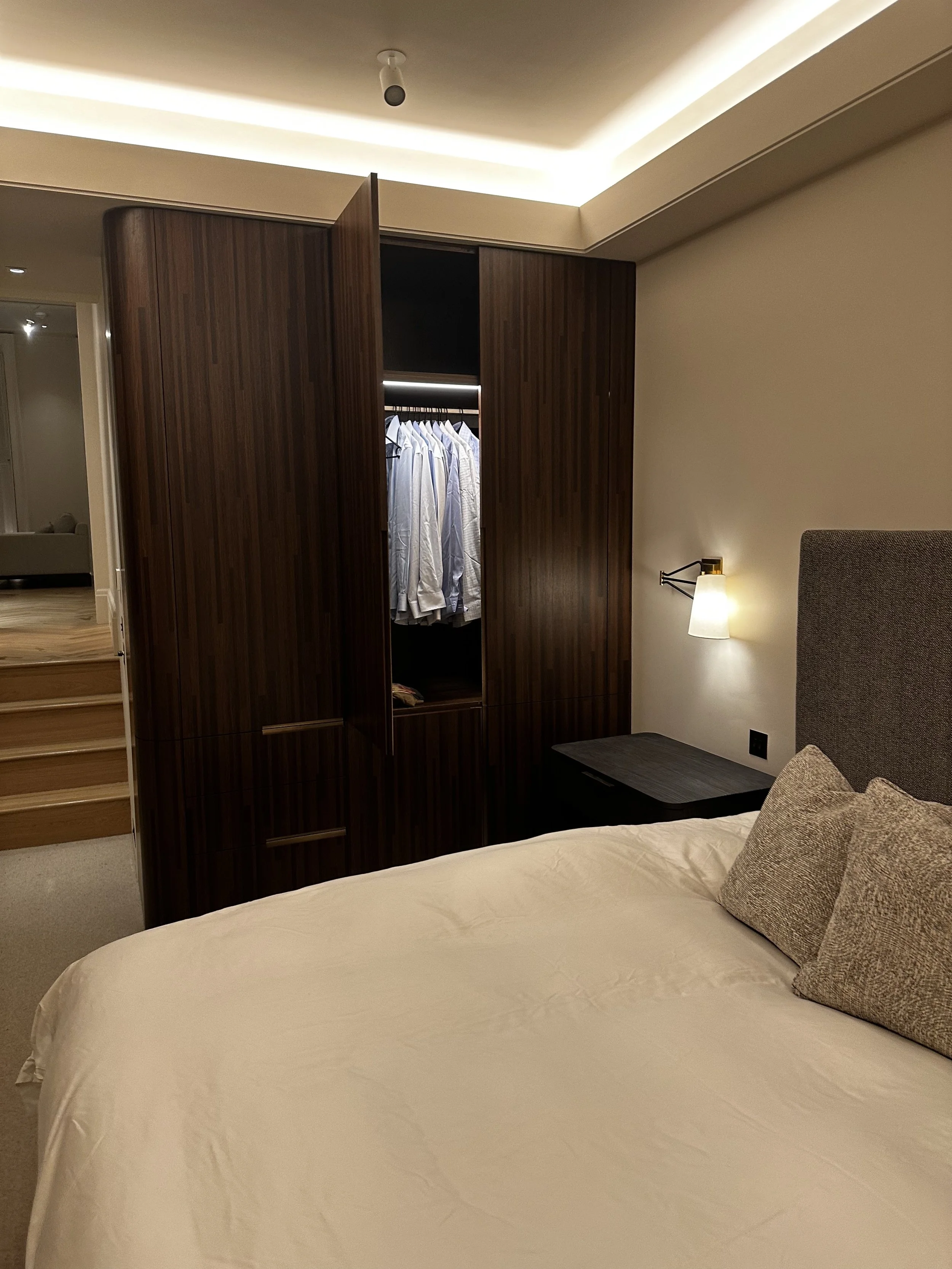 A bedroom with a bed, pillows, a wall-mounted light, a dark wooden nightstand, and an open wardrobe with white shirts hanging inside. Custom bespoke cabinets.