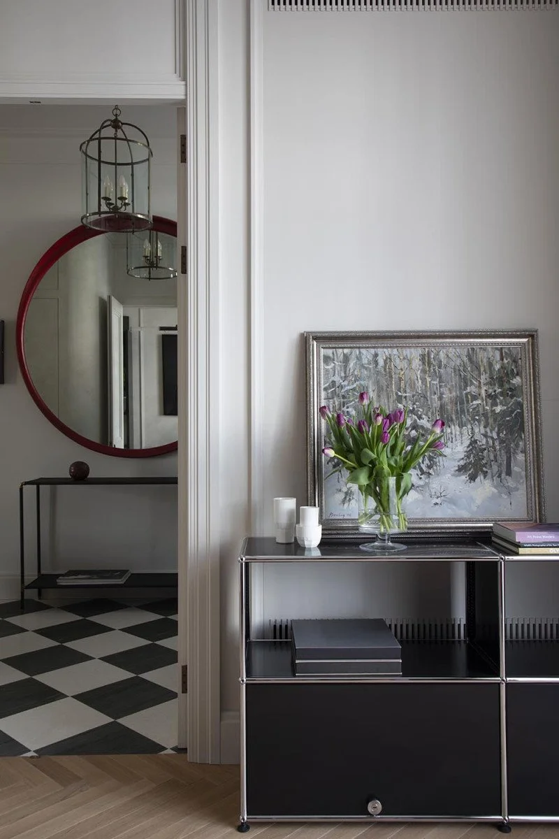 Interior view of a living or hallway space featuring a black and silver console table with a vase of purple tulips, a framed painting of a winter forest, and some candles. The background shows a doorway with black and white checkered flooring and a red-framed mirror.