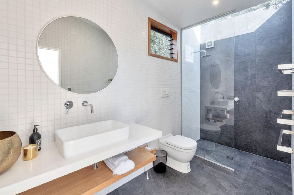 Modern bathroom with white tiled wall, round mirror, vessel sink, toilet, small window, glass shower enclosure with dark gray tiles, and wooden accents.