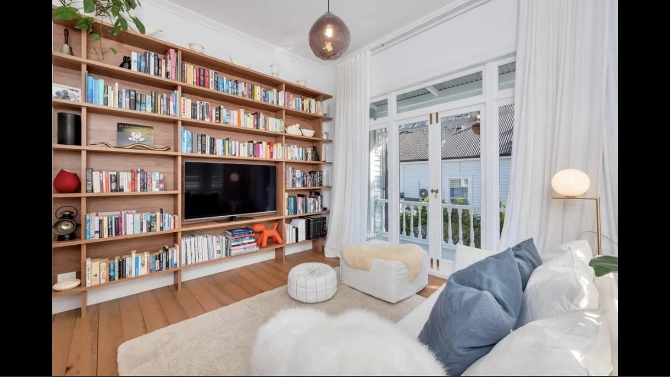 Living room with wooden bookshelf filled with books, a flat-screen TV, a white armchair by a large window, and a white sofa with blue pillows.