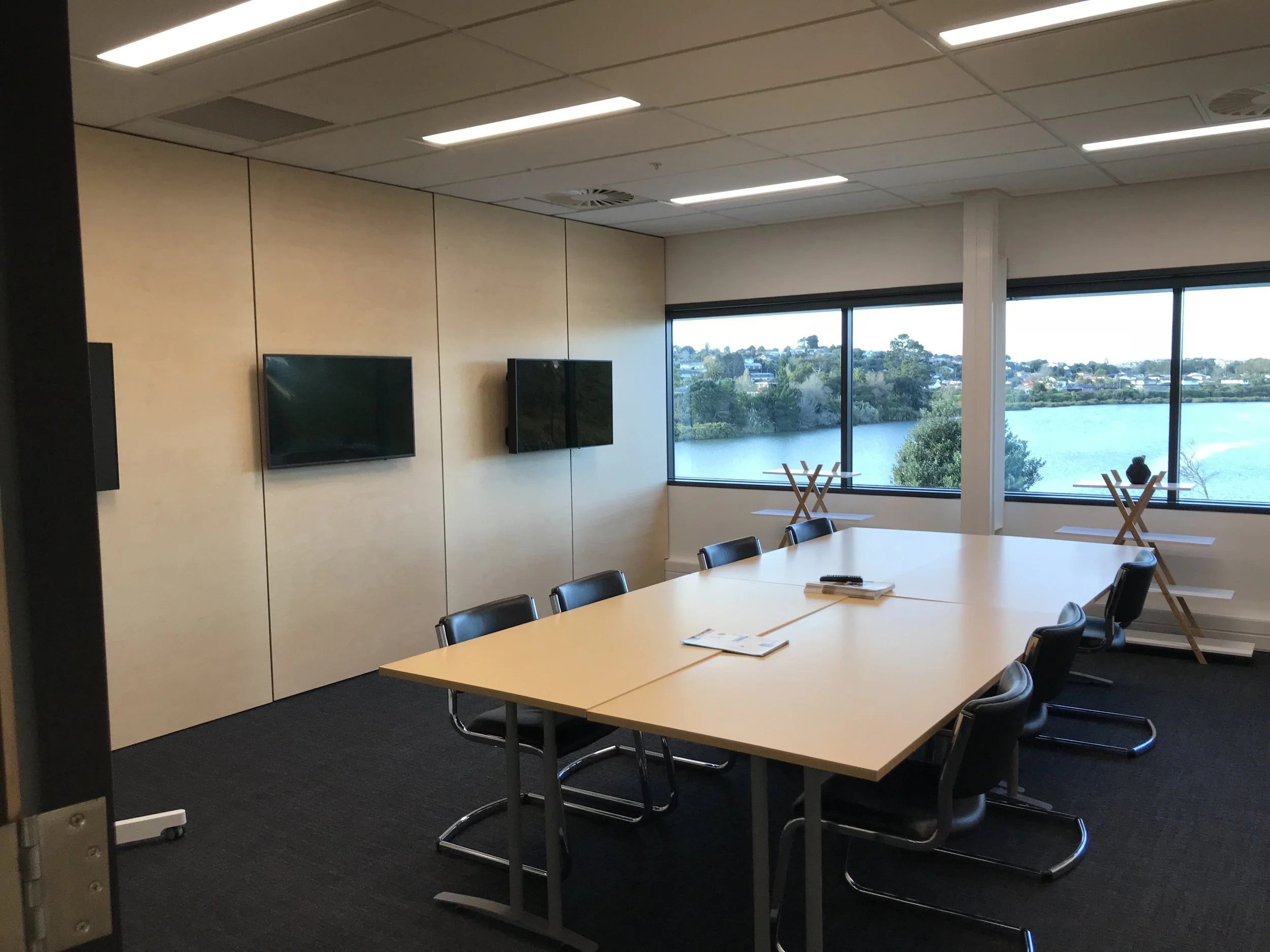 Empty conference room with a long table, black chairs, and large windows overlooking a body of water and greenery.