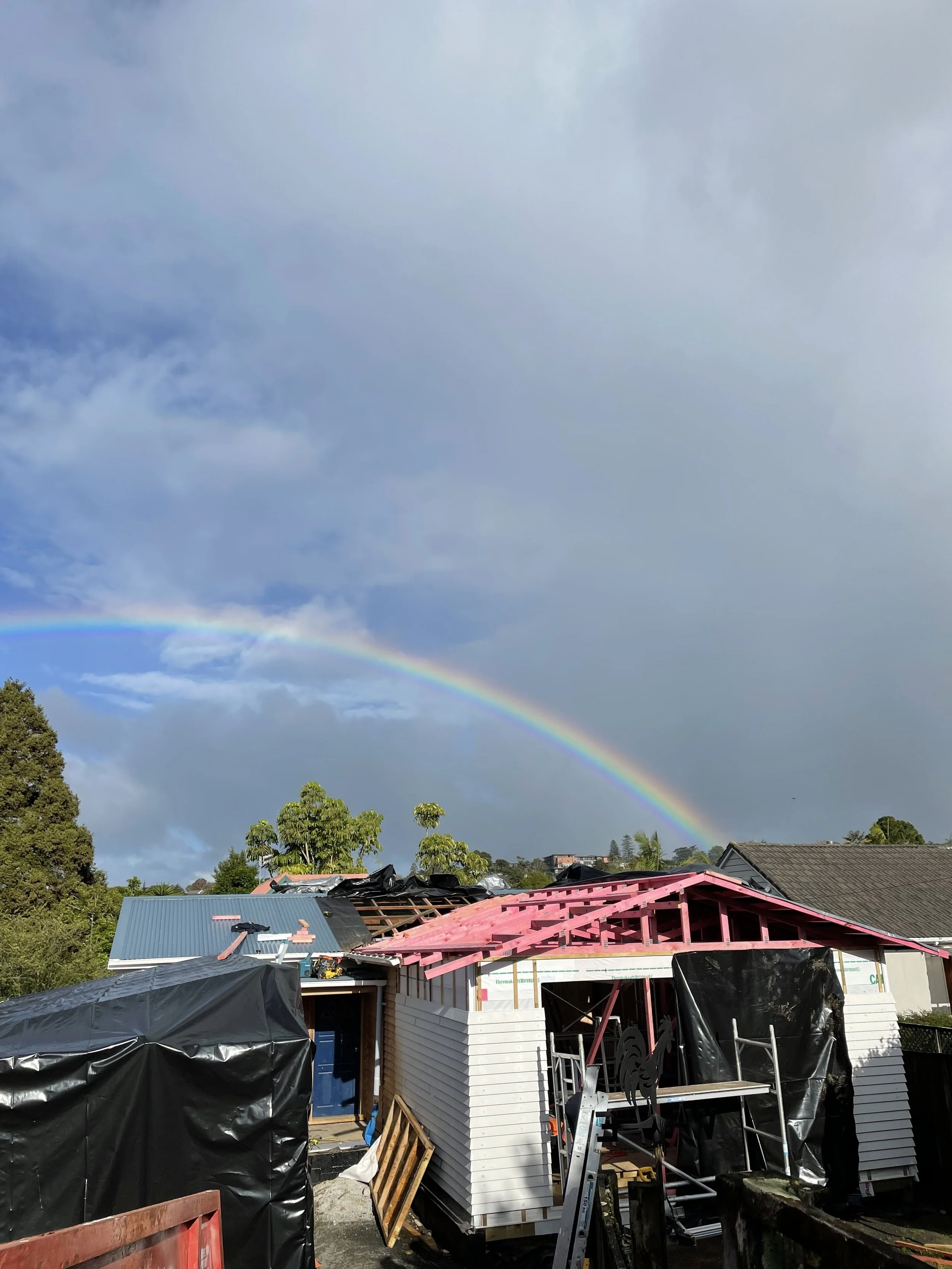 Construction site with a small house being built, featuring pink roof trusses, some covered areas, and a rainbow in the sky.