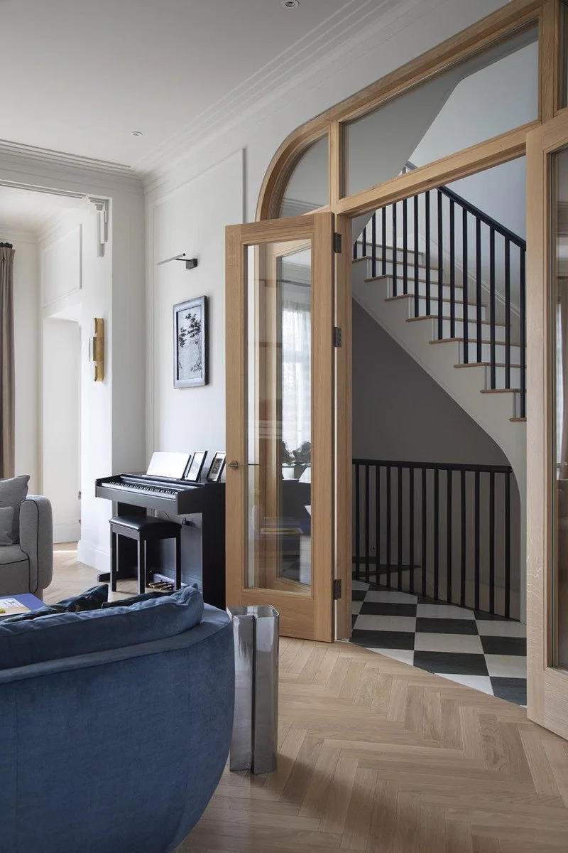Interior view of a living room with a wooden framed glass door leading to a staircase with black railing and checkered black and white floor. There is a furniture with a blue sofa in the foreground, a black digital piano with sheet music on top, and a wall art piece.