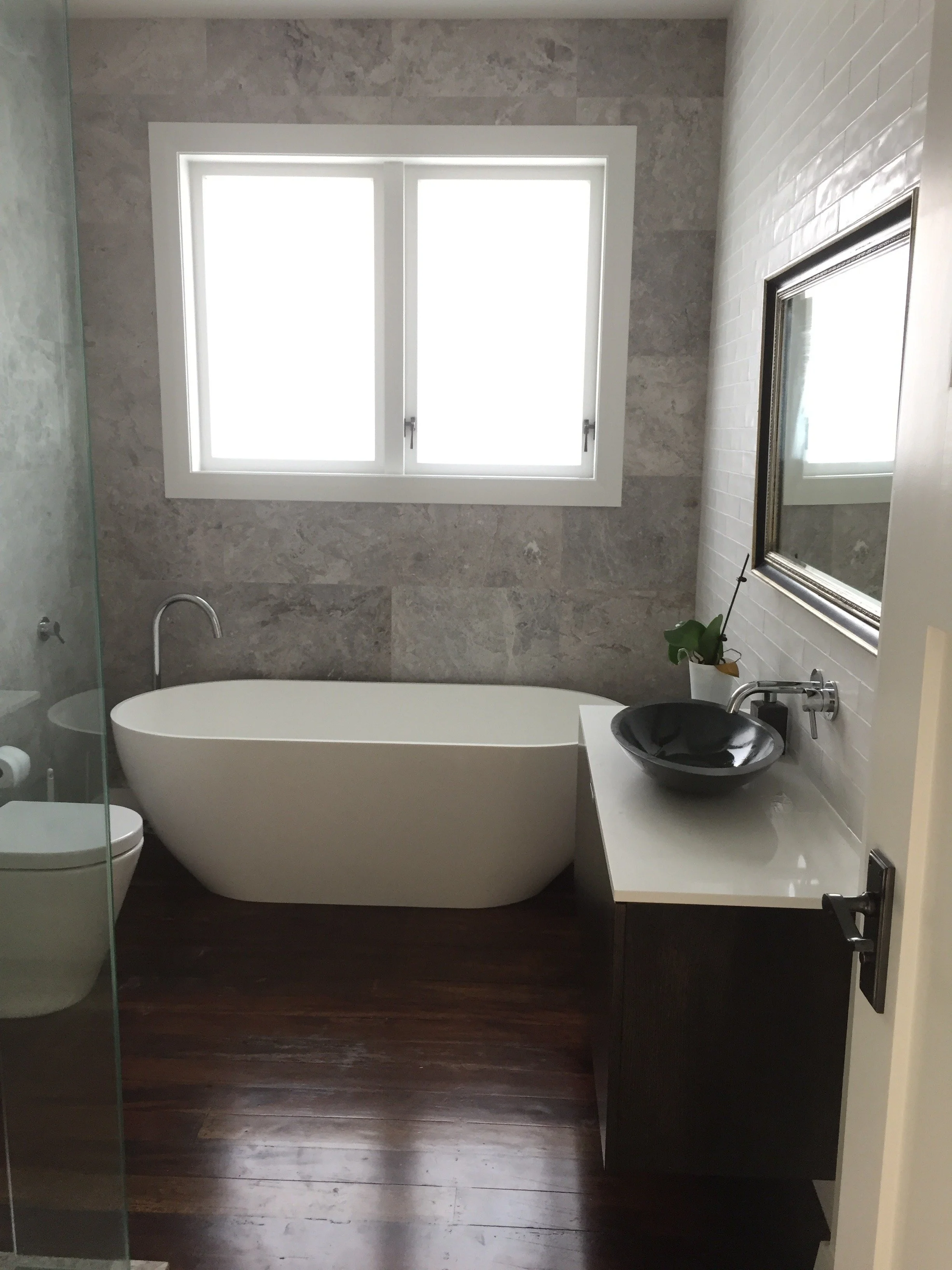 Modern bathroom with a freestanding bathtub, a window above, a black vessel sink on a sleek white countertop, and a mirror on the wall.