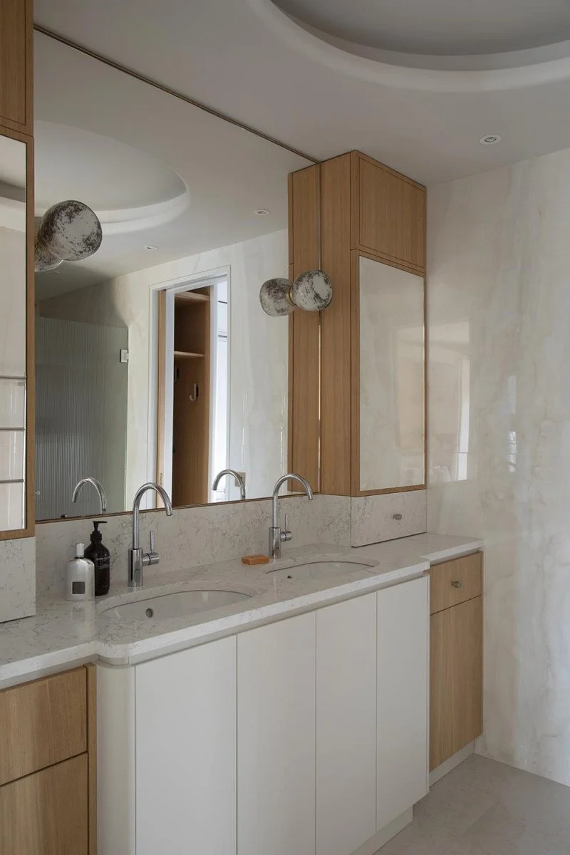 A modern bathroom vanity with a marble countertop, double sinks, wooden cabinet doors, and large mirror, with decorative pendant lights and toiletries on the counter.