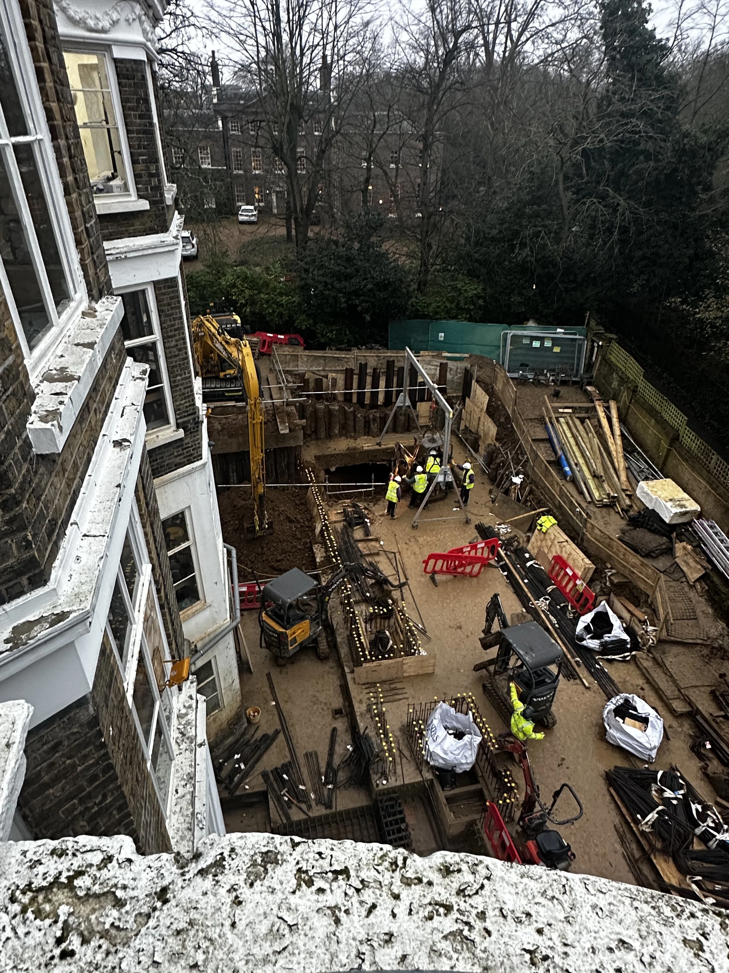 Construction workers at a site next to a building, surrounded by equipment, safety barriers, and materials, with trees and parked cars in the background.