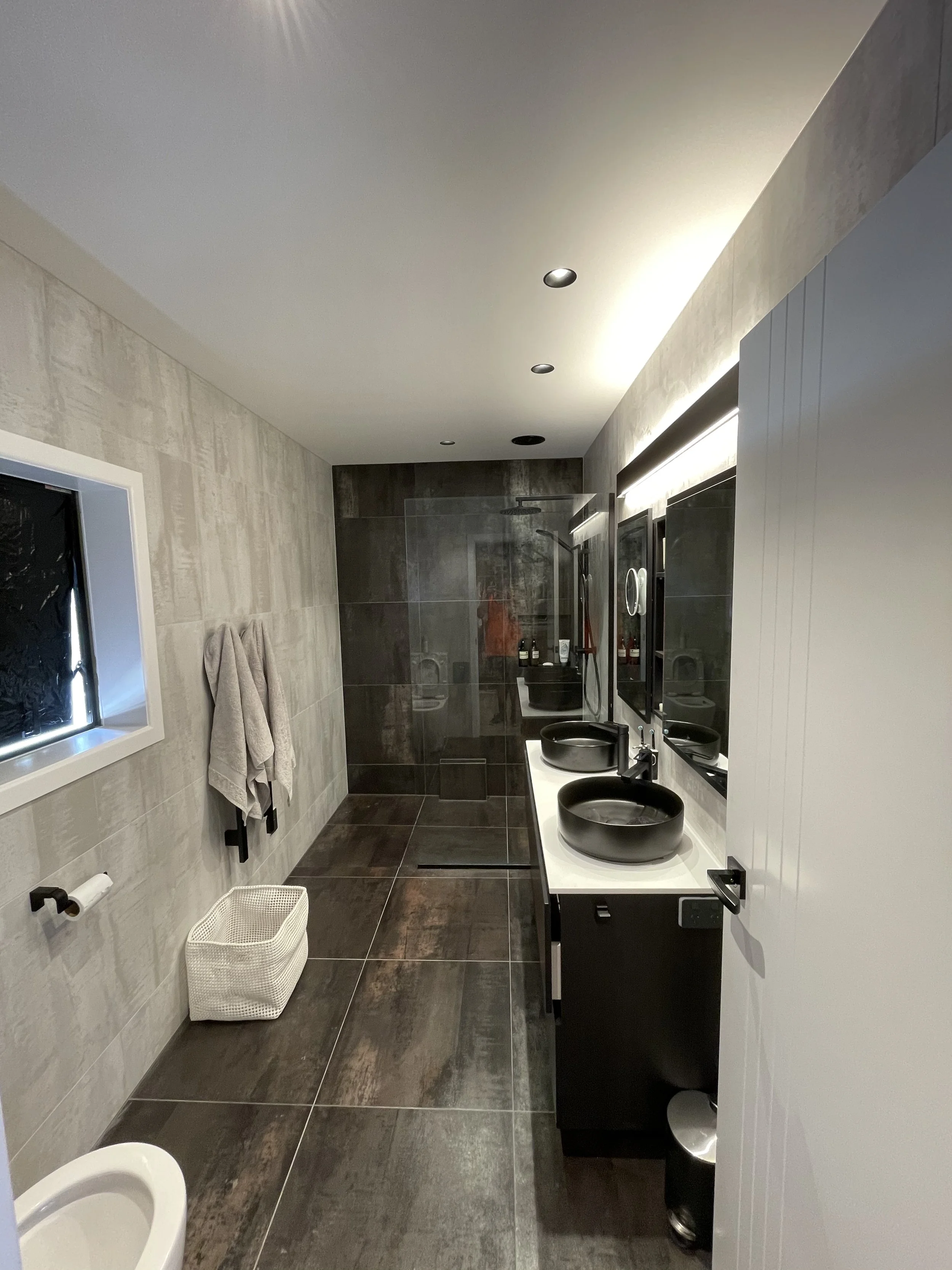 Modern bathroom with dark tiled floor and walls, featuring two black vessel sinks on a white countertop, mirrors, and a walk-in shower with glass enclosure at the back.