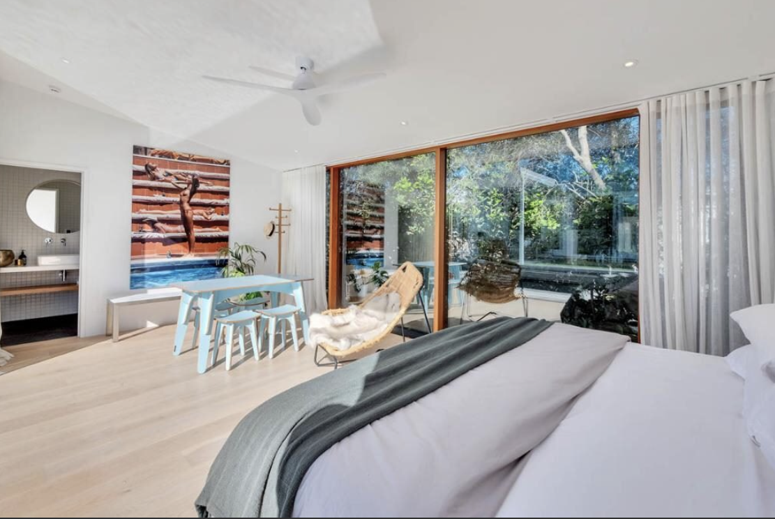 Bright hotel room with large sliding glass doors, a bed with gray and white bedding, a wicker chair with a blanket, a minimalist dining table with blue chairs, and a view of trees outside