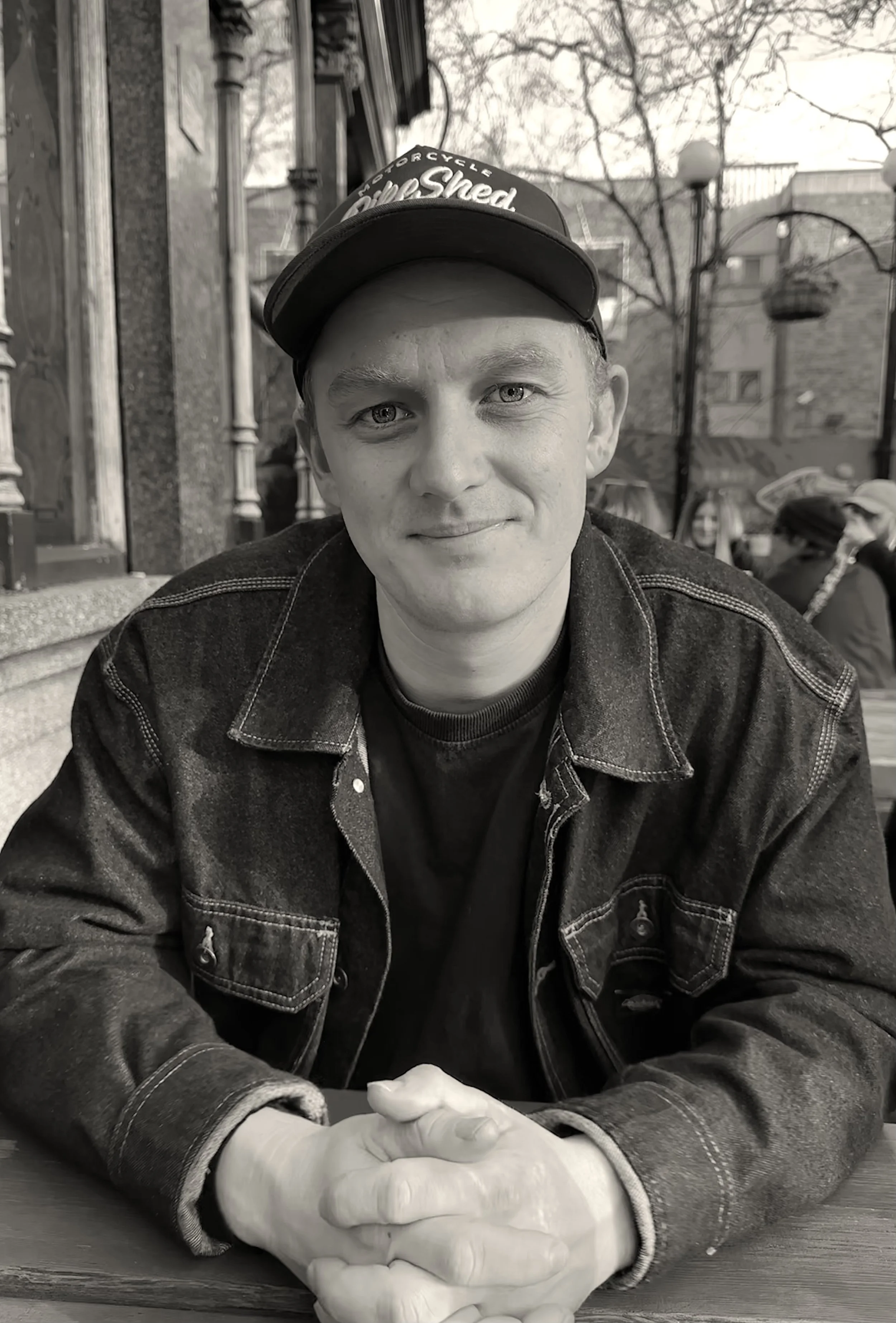 A young man sitting at a table outdoors, wearing a black cap and denim jacket, smiling at the camera, with a background of outdoor furniture, trees, and buildings in black and white.