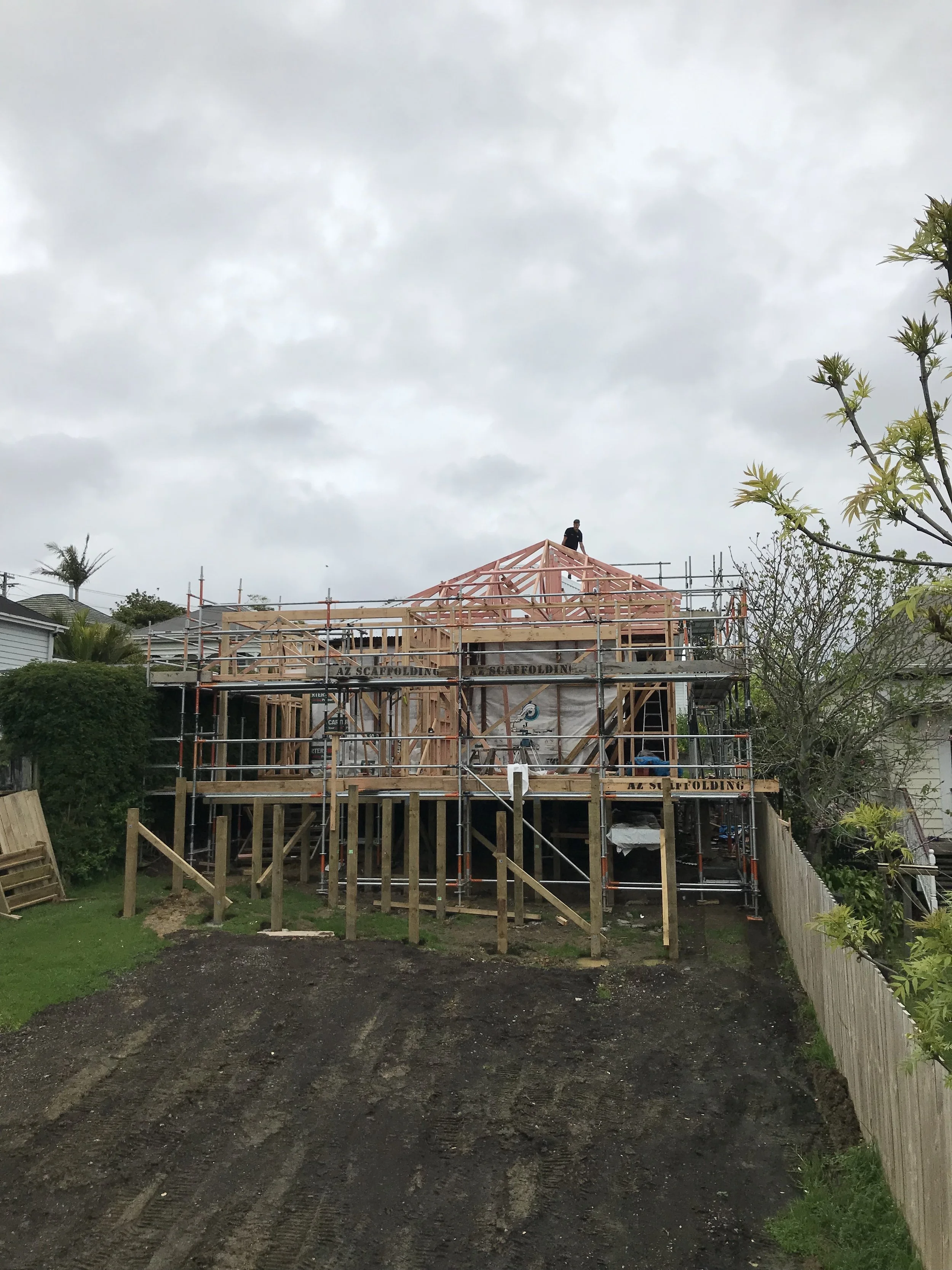 A house under construction with wooden framing, scaffolding, and a worker on top of the roof frame. The yard has some grass, dirt, and a fence on the right side.