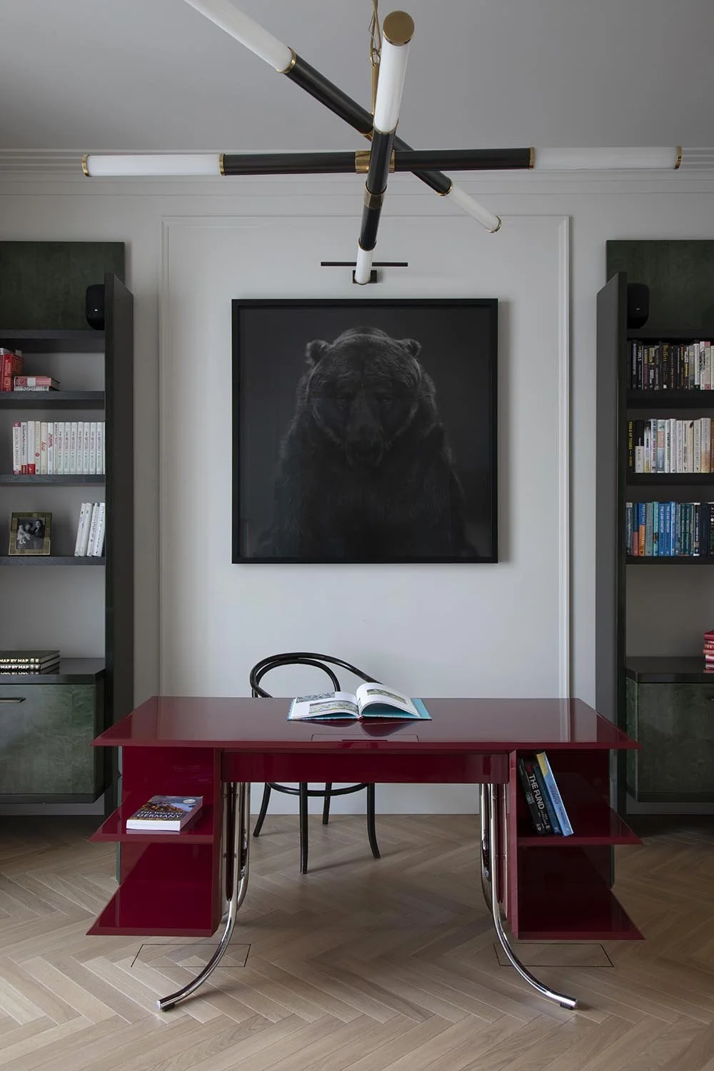 A home office with a red desk, black chair, bookshelf on each side, black and white bear portrait on the wall, open book on desk, and modern black and brass ceiling light fixture. Expert wooden floor detailing.