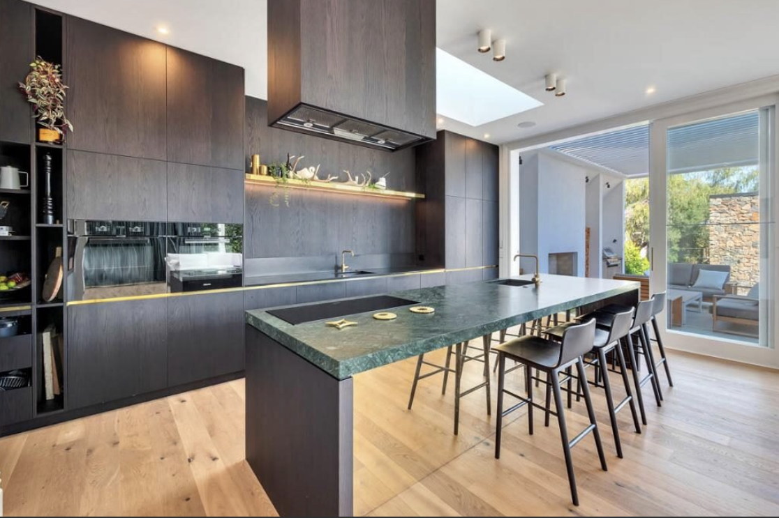 Modern kitchen with dark wood cabinets and a green marble island, bar stools, and large windows with outdoor seating.