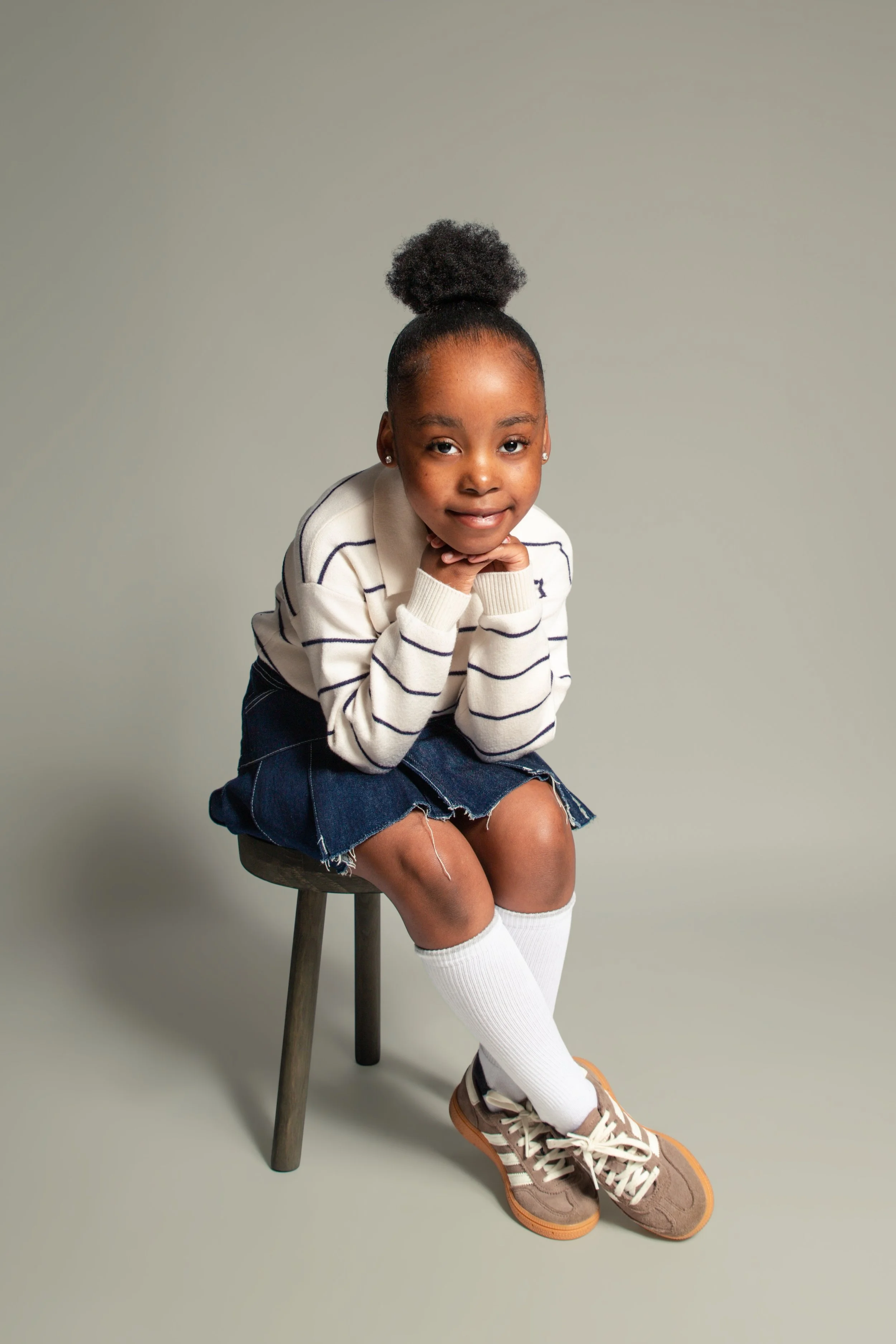 young girl sat on a stool with her hands under her chin looking at camera. Feet crossed over for an at home children's portrait photoshoot in Bury