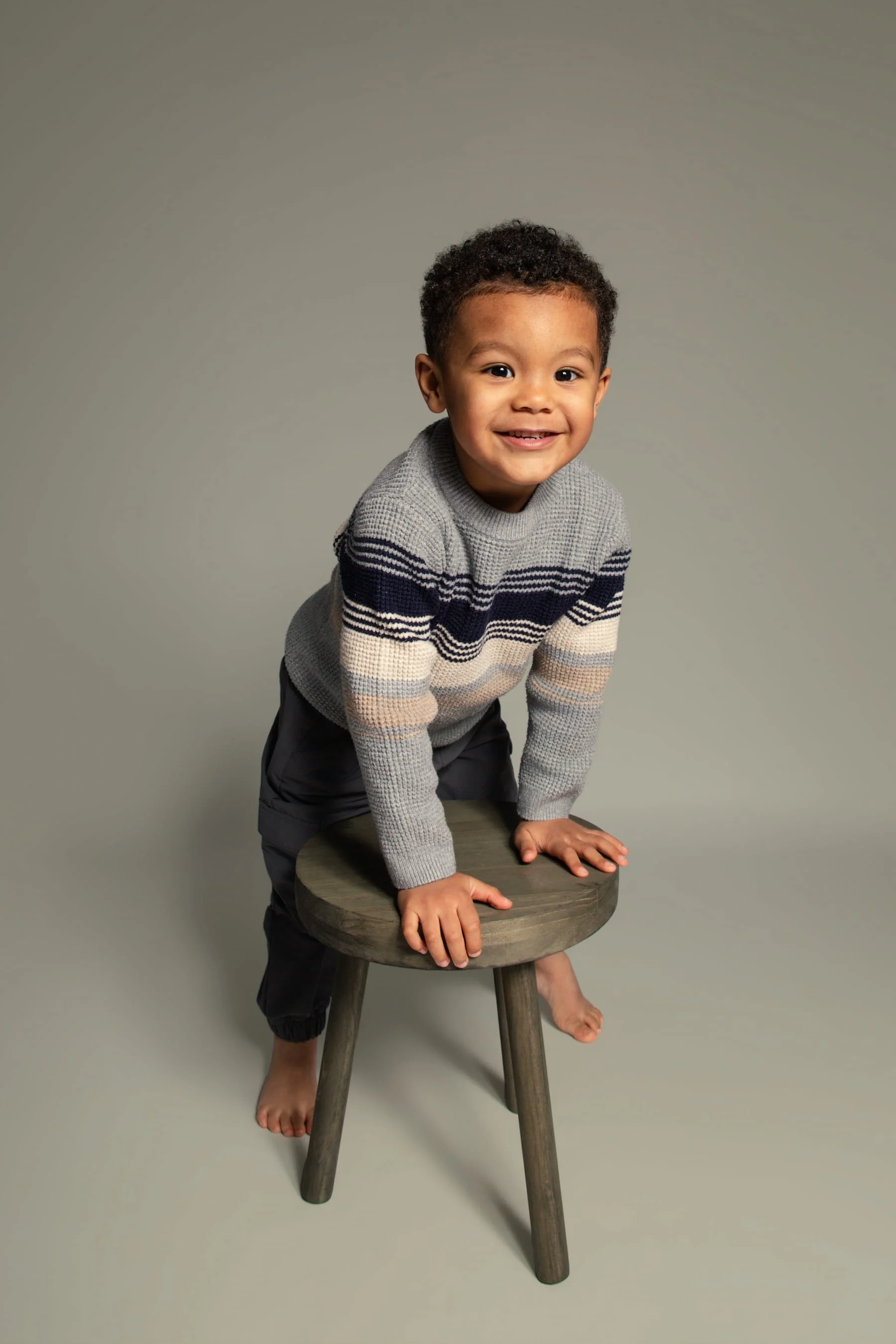 Natural at-home portrait of young boy leaning on a stool and smiling, captured in a relaxed and playful style.