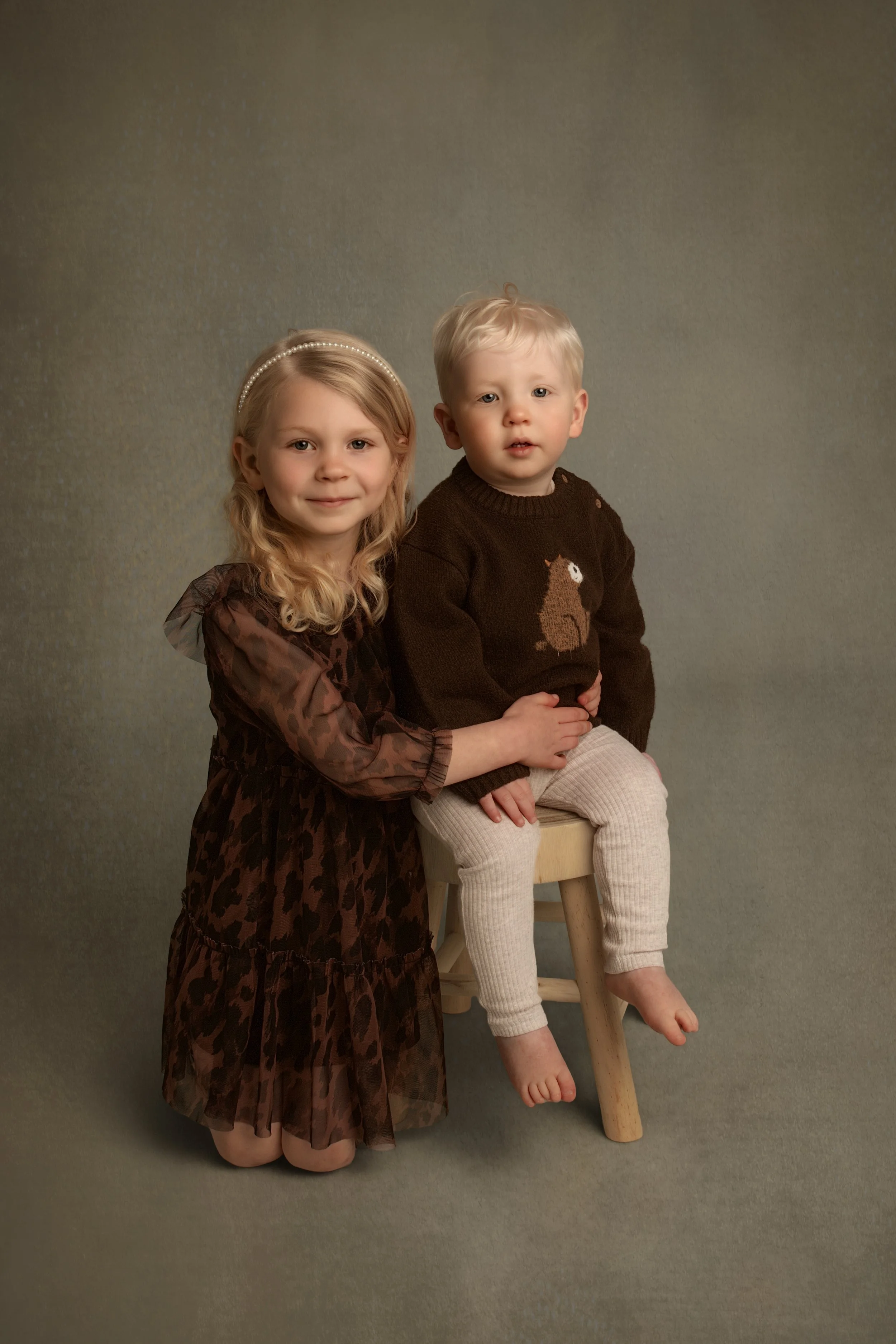 Fine art sibling portrait of young girl kneeling beside her toddler brother seated on a stool, captured using soft studio lighting in a timeless fine art style.