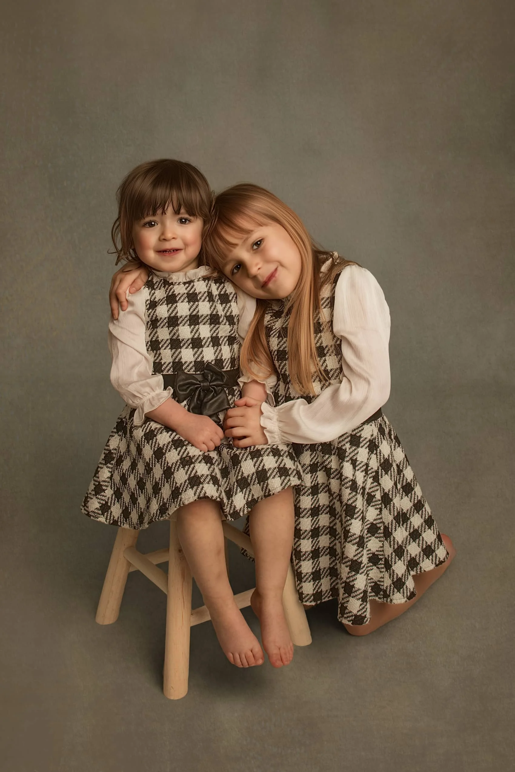 Fine art sibling portrait of two young sisters, with toddler seated on a stool, styled in a timeless fine art style.