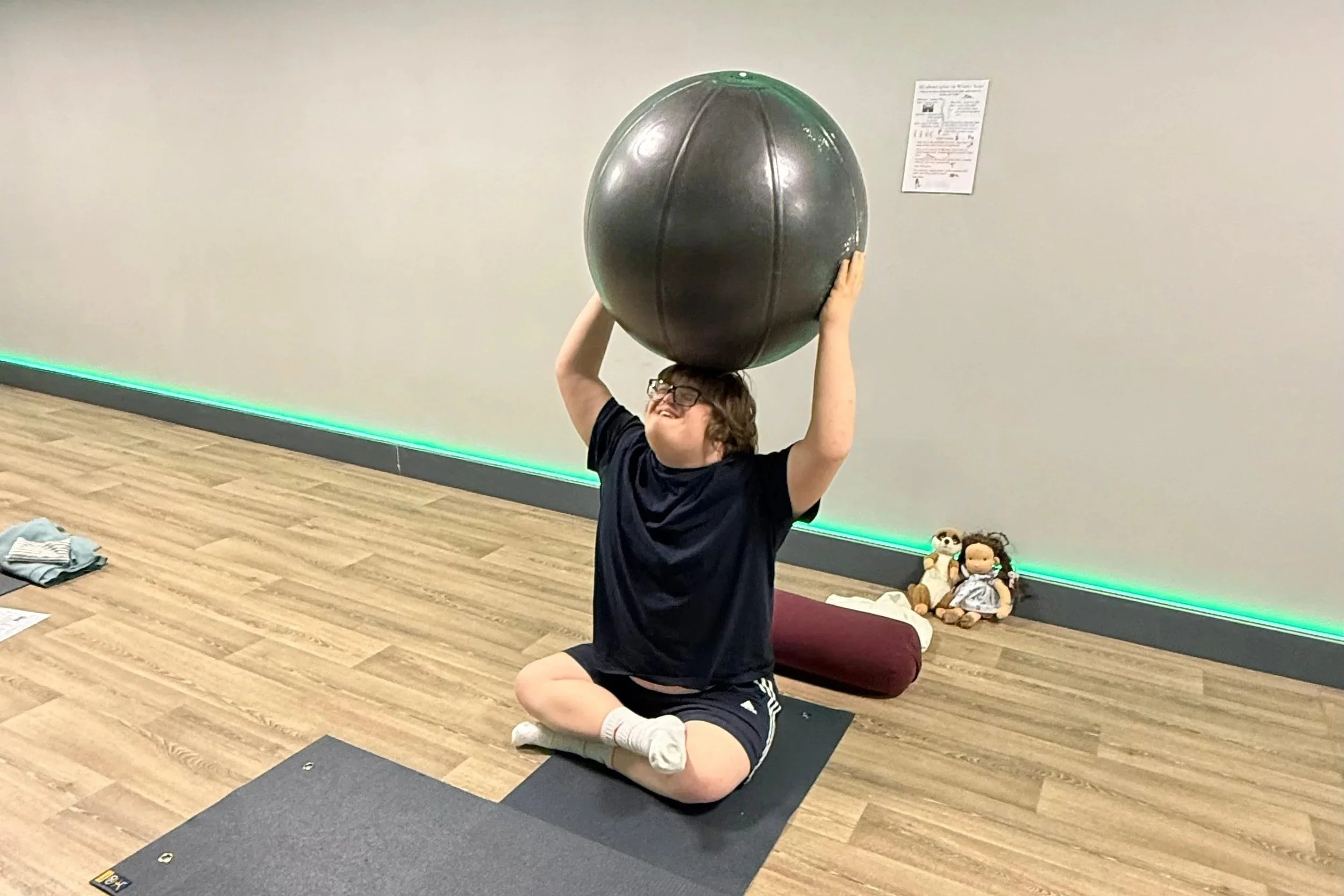 Person sitting cross-legged on a yoga mat, holding a large exercise ball above their head in a gym or fitness studio with wood flooring.