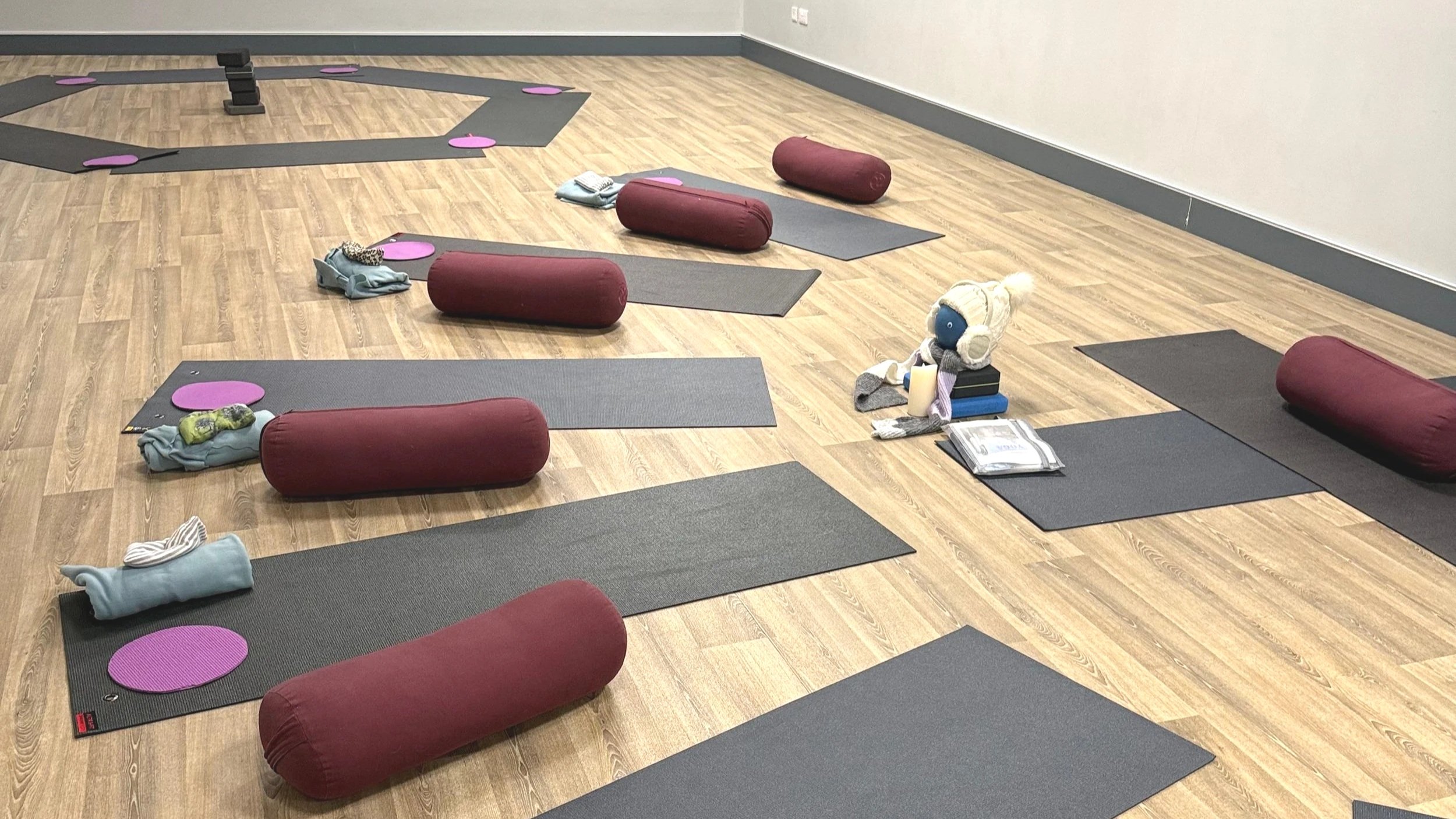 Yoga studio with black mats, pink meditation cushions, and various items on the floor, including folded clothes, a stuffed animal, a book, and a plastic bag, in a room with wooden floors and plain walls.