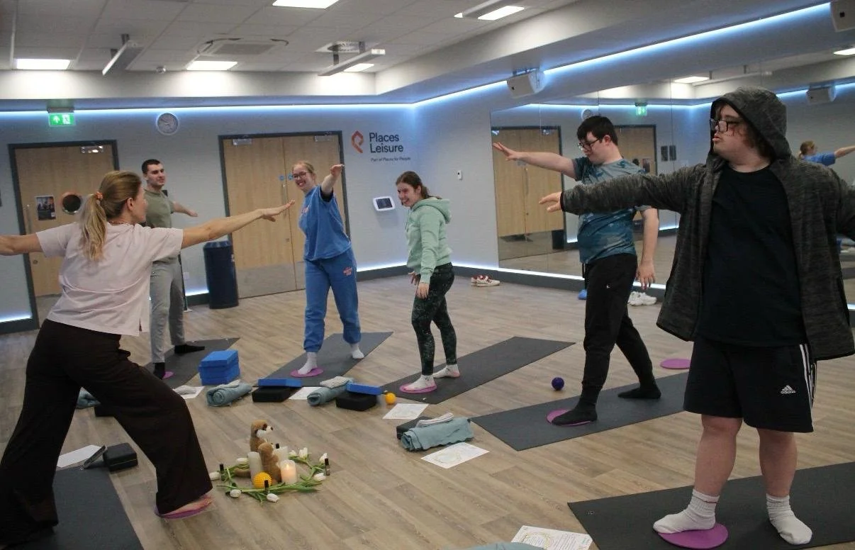 A group of people participating in a yoga or stretching class in a fitness studio, with mats and yoga props on the floor, and a mirror reflecting the scene.