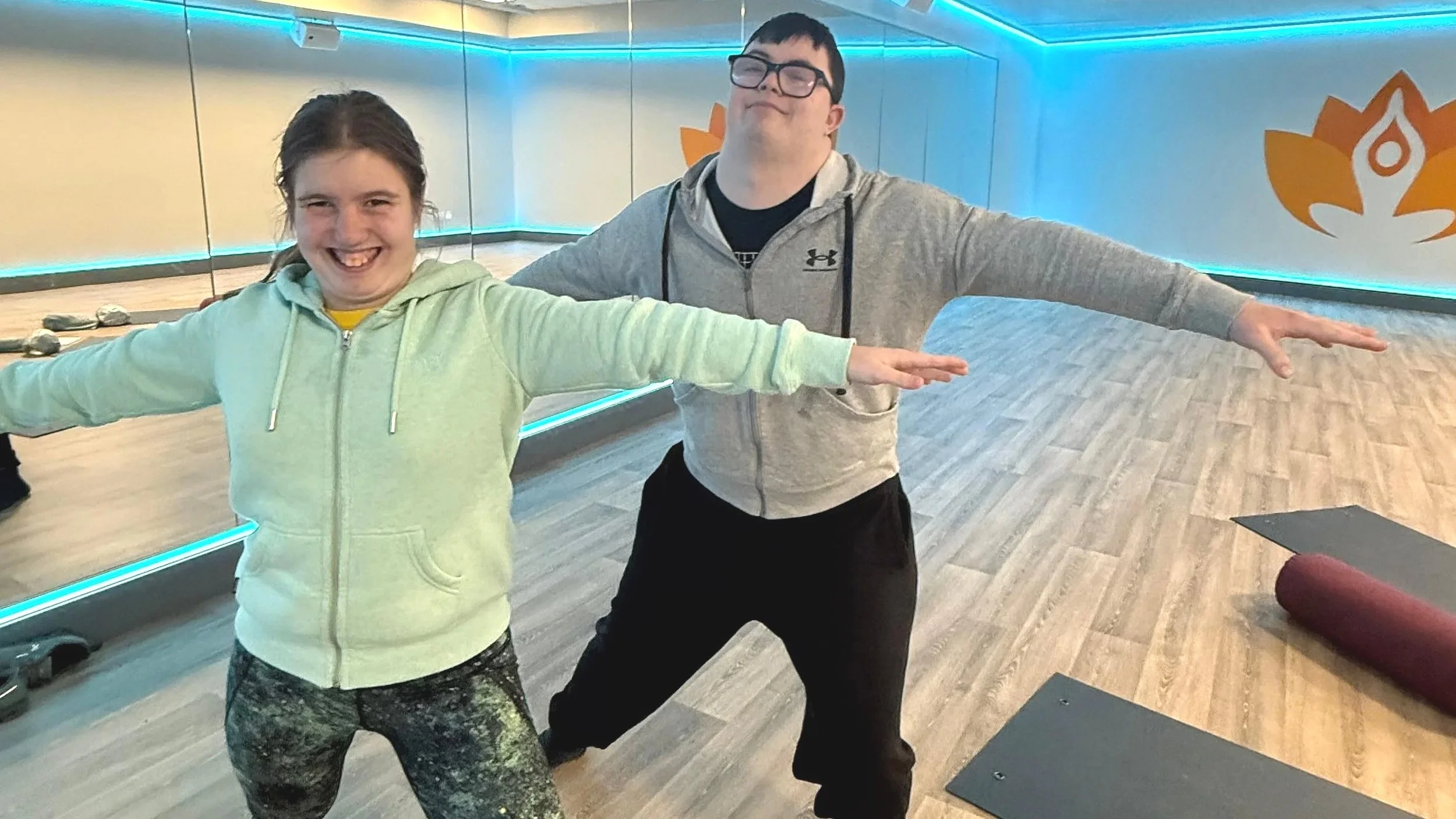 A young girl and a man standing in a yoga studio with arms extended to the sides, smiling and posing for the camera. The studio has wooden floors, a large mirror, and blue lighting.