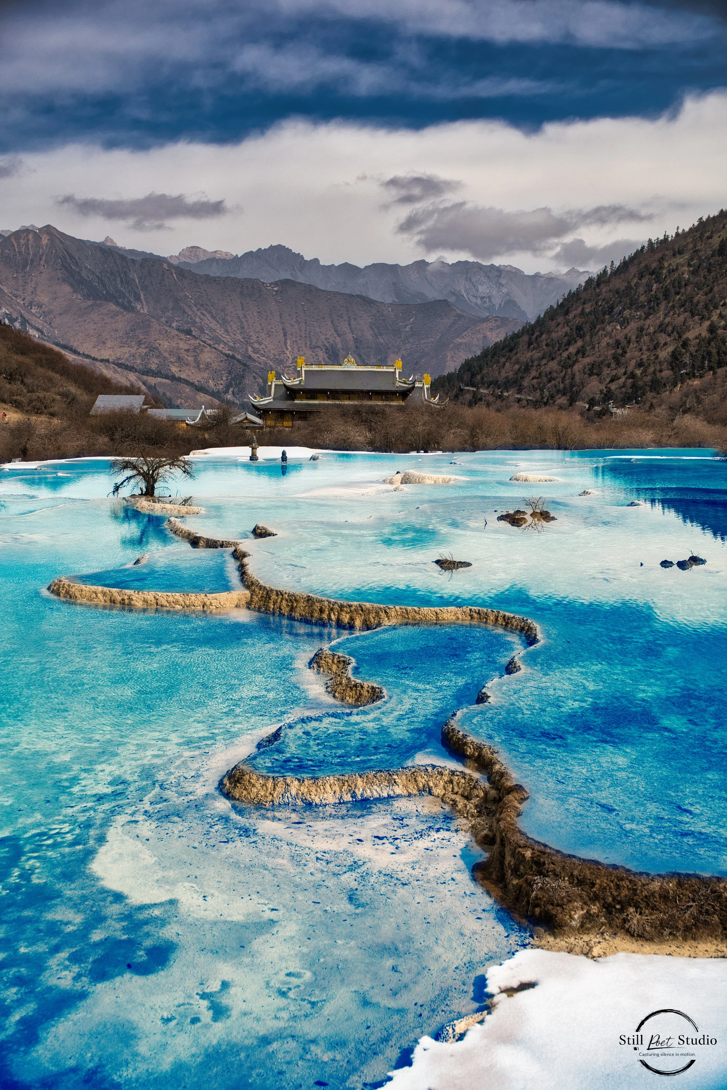 Scenic landscape of terraced hot springs with bright blue mineral-rich water, traditional Asian-style building in the distance, mountains, and cloudy sky in the background.