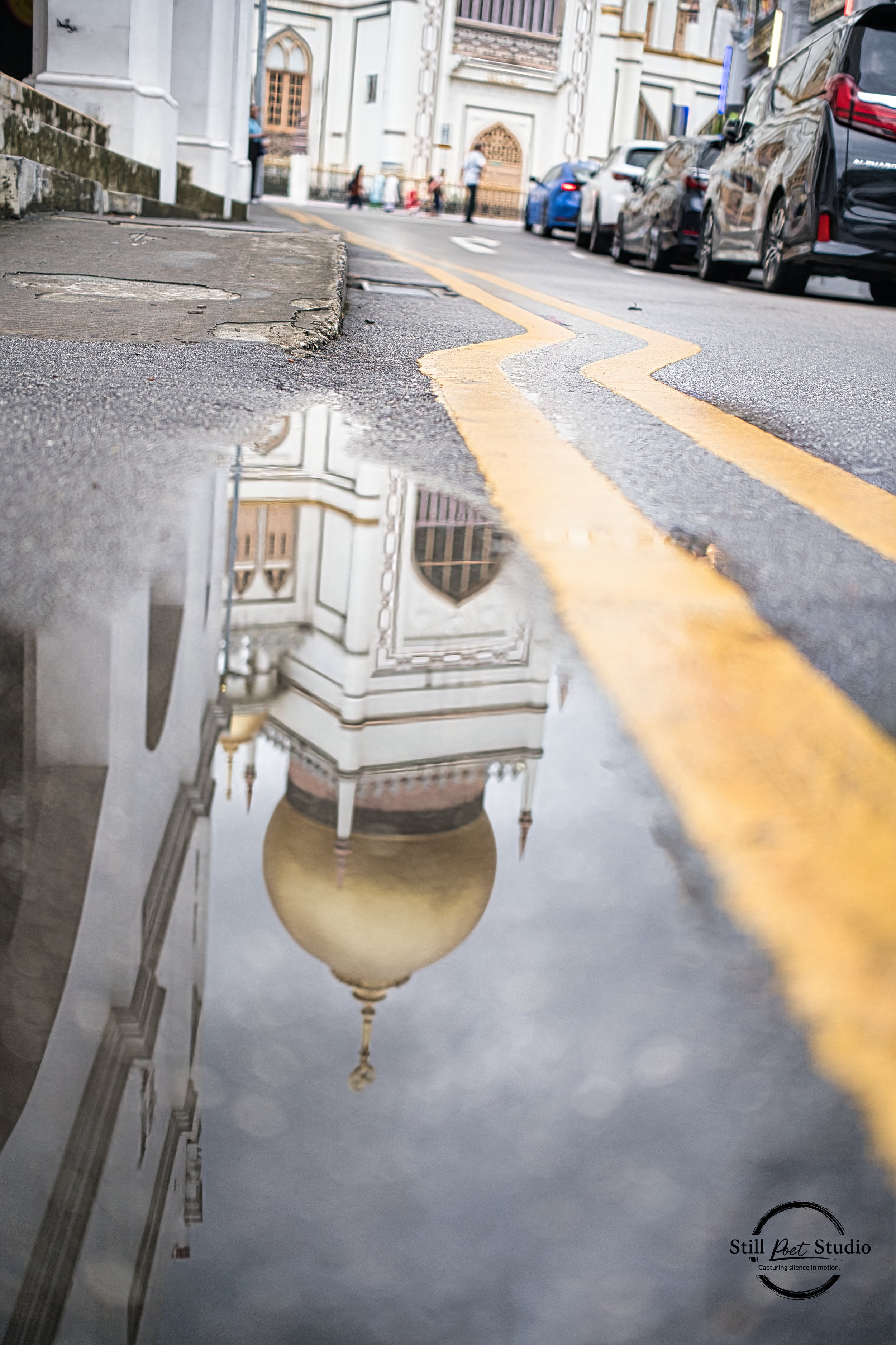 Reflection of a white building with a domed tower in a puddle on a city street, with parked cars and pedestrians in the background.