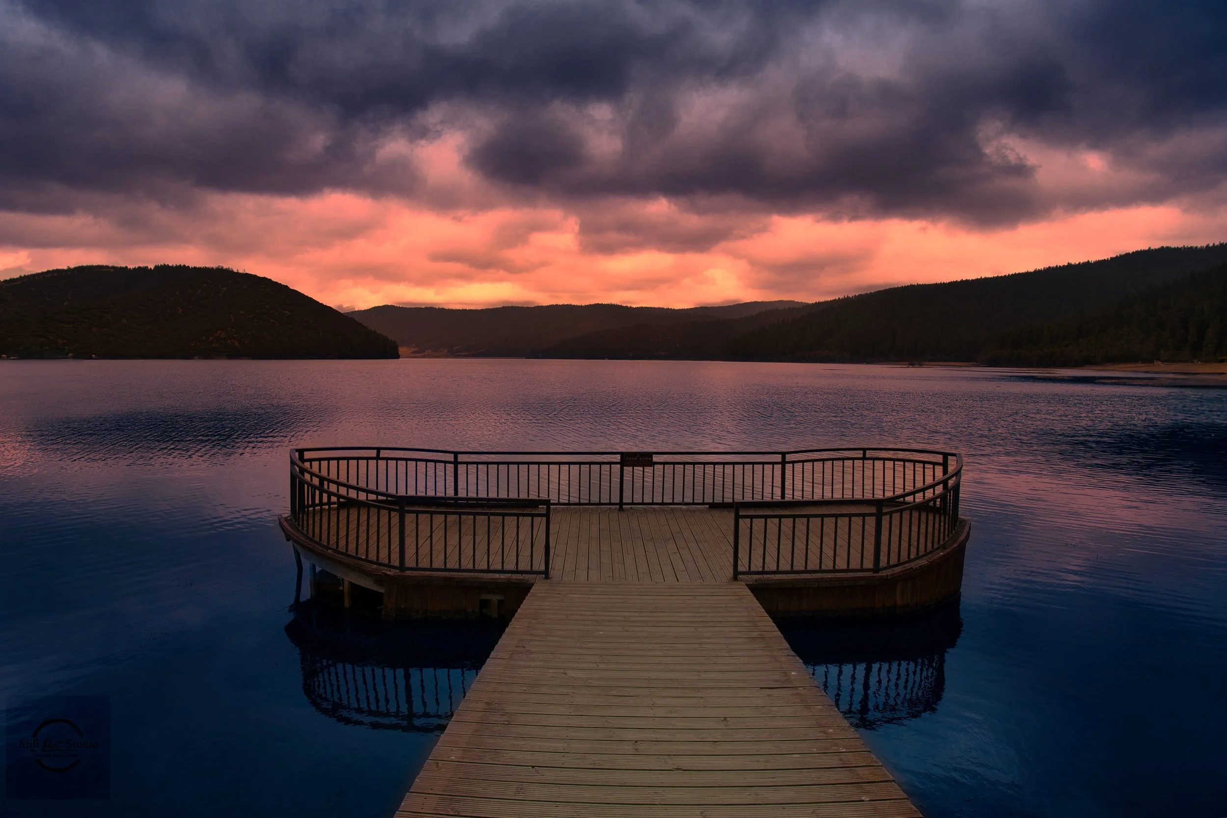 A wooden dock with a curved railing extends into a calm lake during sunset, with dark clouds and pinkish purple hues in the sky, surrounded by distant hills covered in dark trees.