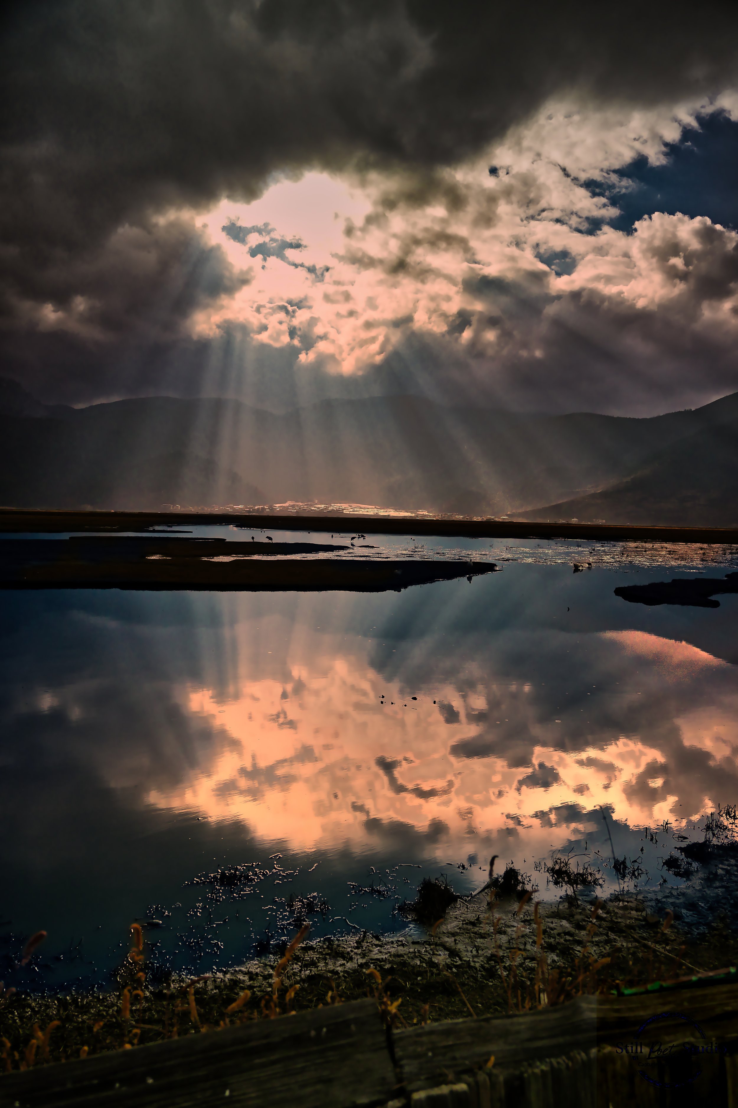 Sunlight beams breaking through dark clouds over mountains and a body of water, creating reflections of the sky and clouds on the water surface.