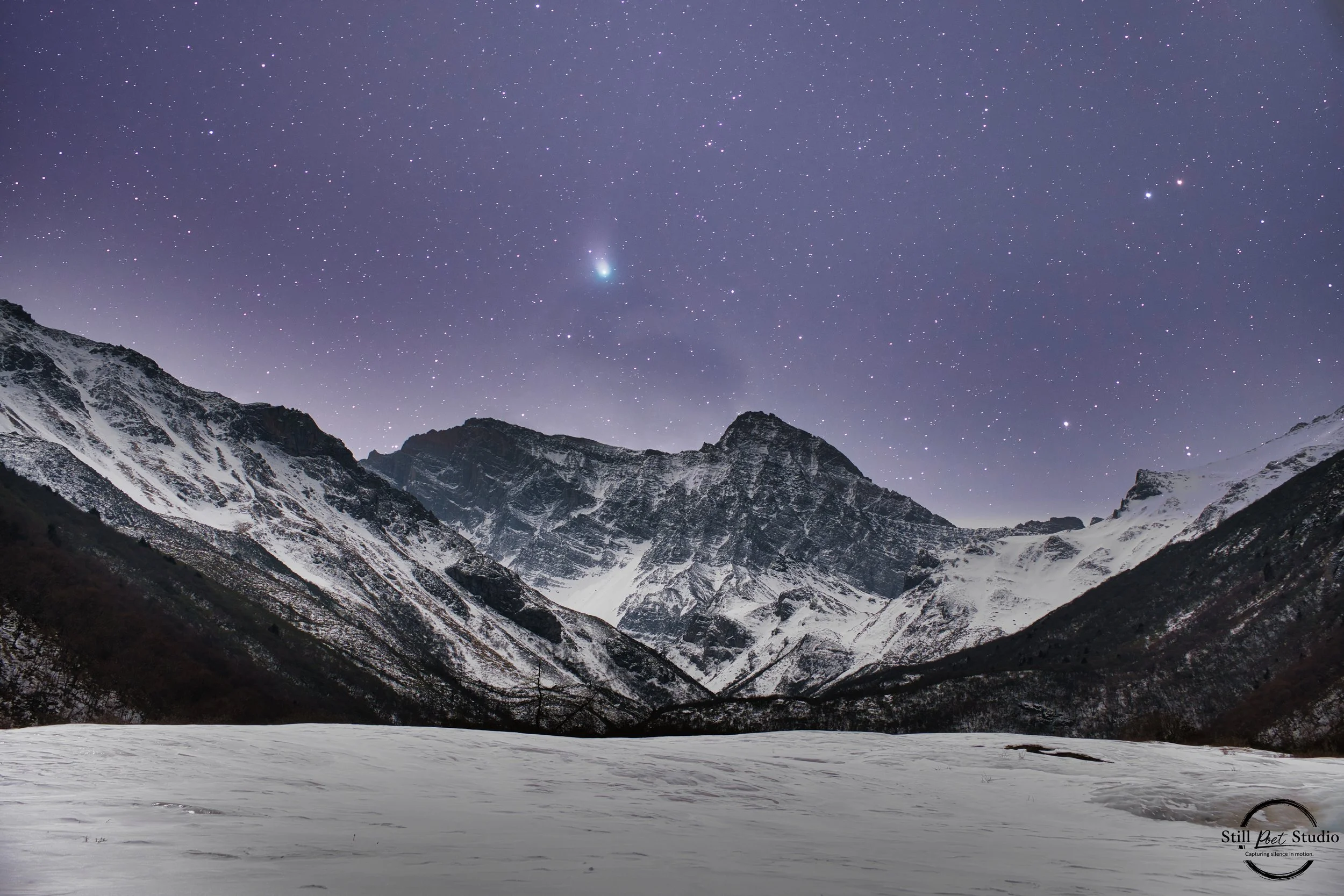 Snow-covered mountain landscape under a starry night sky with a visible comet and numerous stars.