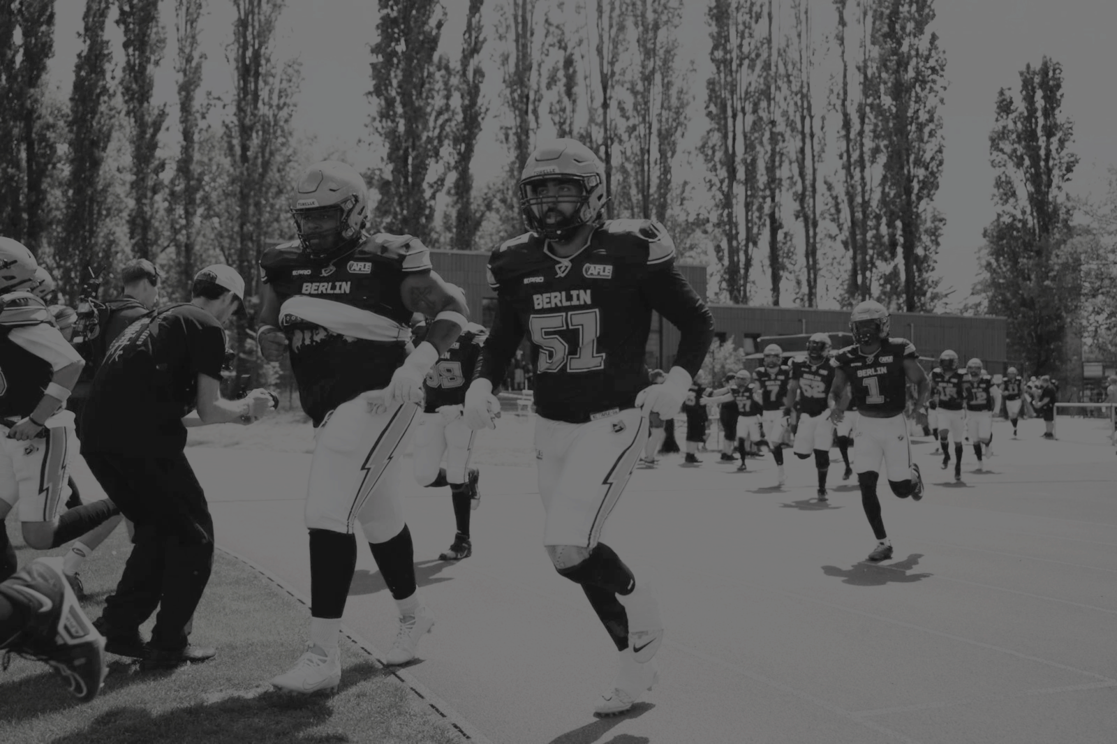 American-Footballspieler in schwarzen Trikots, die auf dem Spielfeld im Freien laufen, mit Trainern und anderen Spielern im Hintergrund.