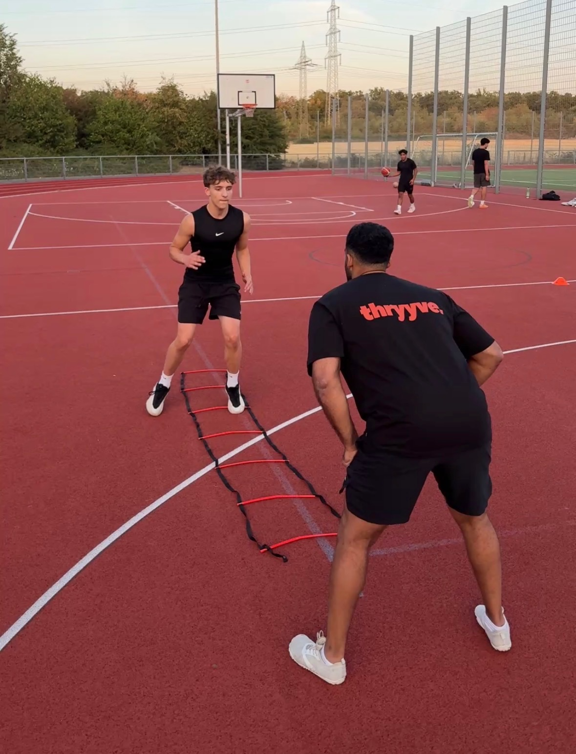 Jugendlicher trainiert auf einem Tennisplatz mit einem Koordinationsleiter, überwacht von einem Trainer, während im Hintergrund zwei weitere Jugendliche mit Basketbällen üben.