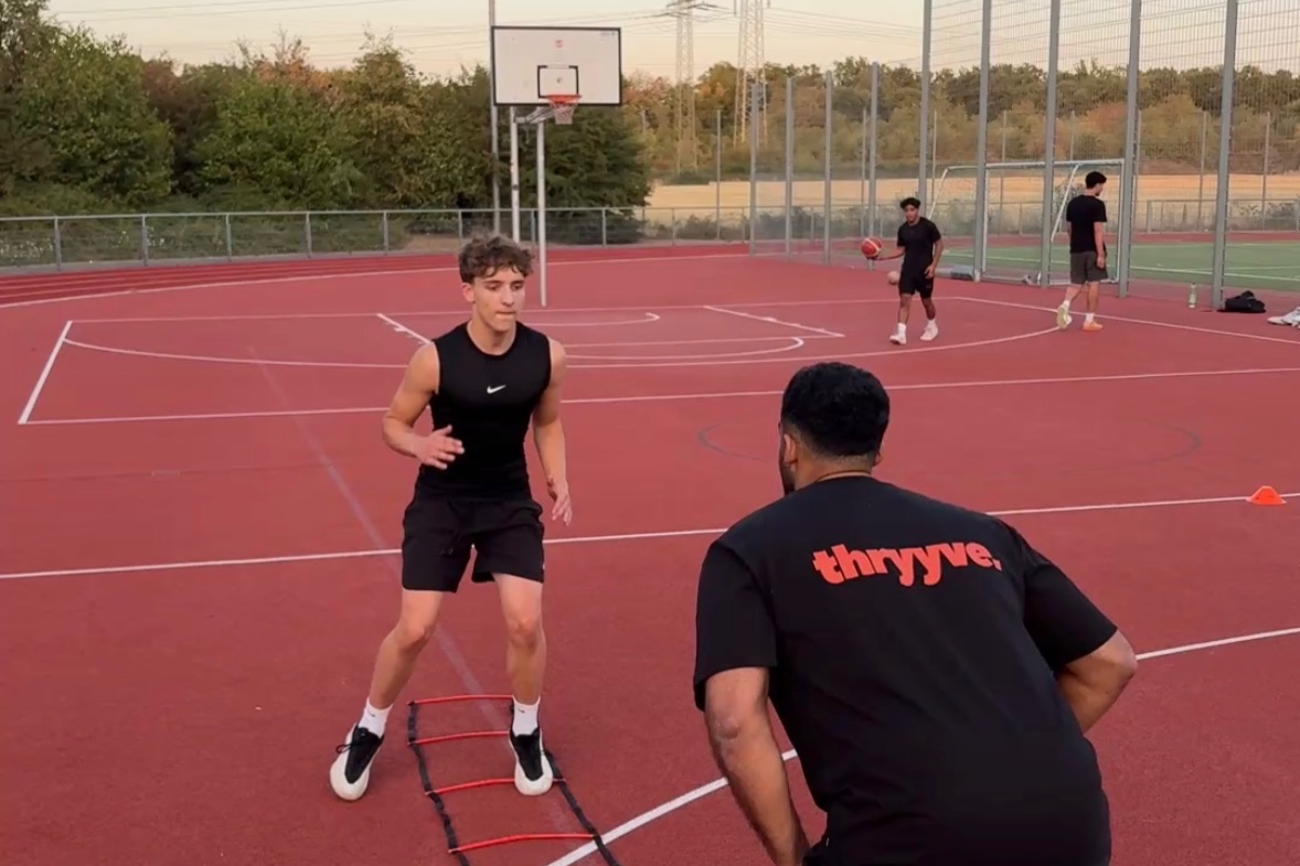 Ein junger Mann trainiert auf einem roten Tennisplatz mit einem Schlagschnur, während ein Trainer ihn überwacht. Im Hintergrund sind andere junge Männer beim Basketball spielen.