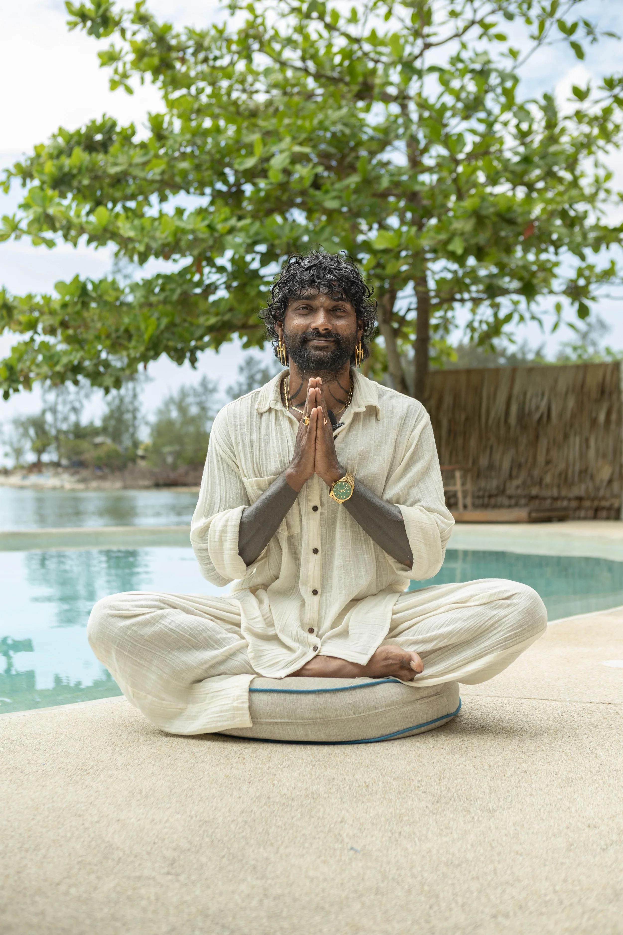 A man with curly hair, beard, and jewelry sitting cross-legged in a yoga pose outdoors near a body of water, with trees and a thatched structure in the background, under a cloudy sky.