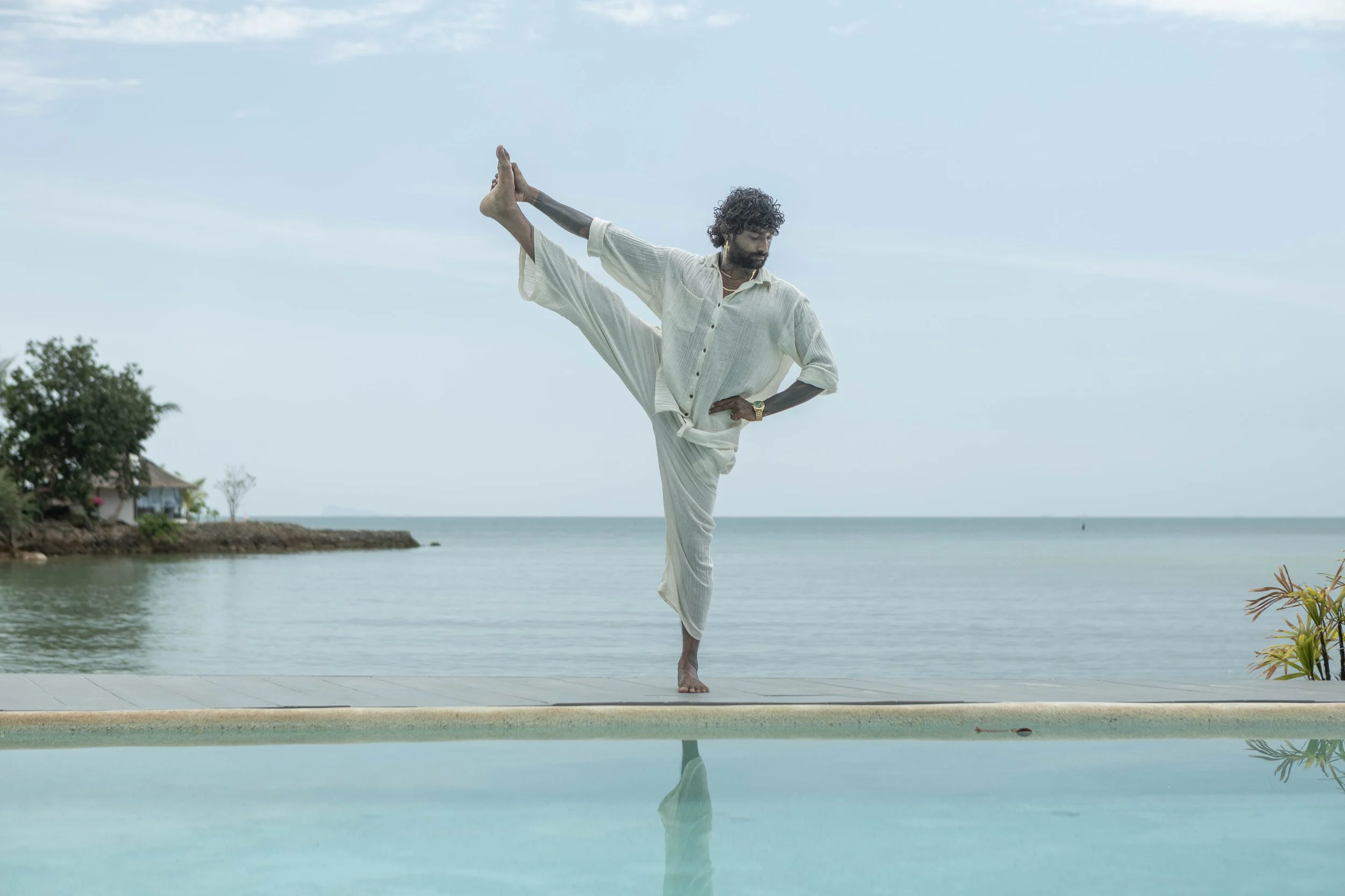 A man in traditional Indian attire practicing yoga by a swimming pool on the beach at sunset.
