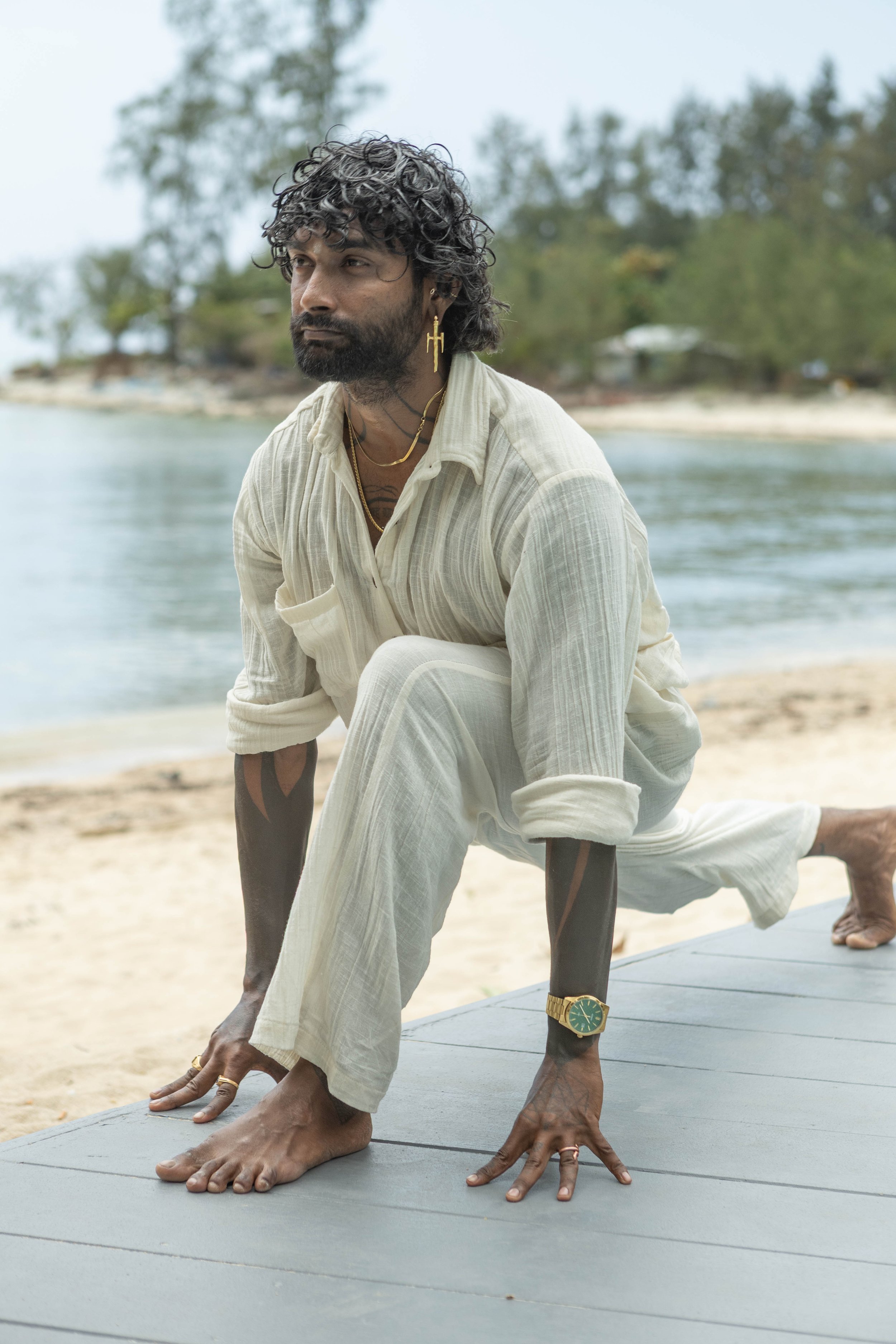 Man practicing yoga outdoors on a wooden platform near a body of water, with trees and cloudy sky in the background.